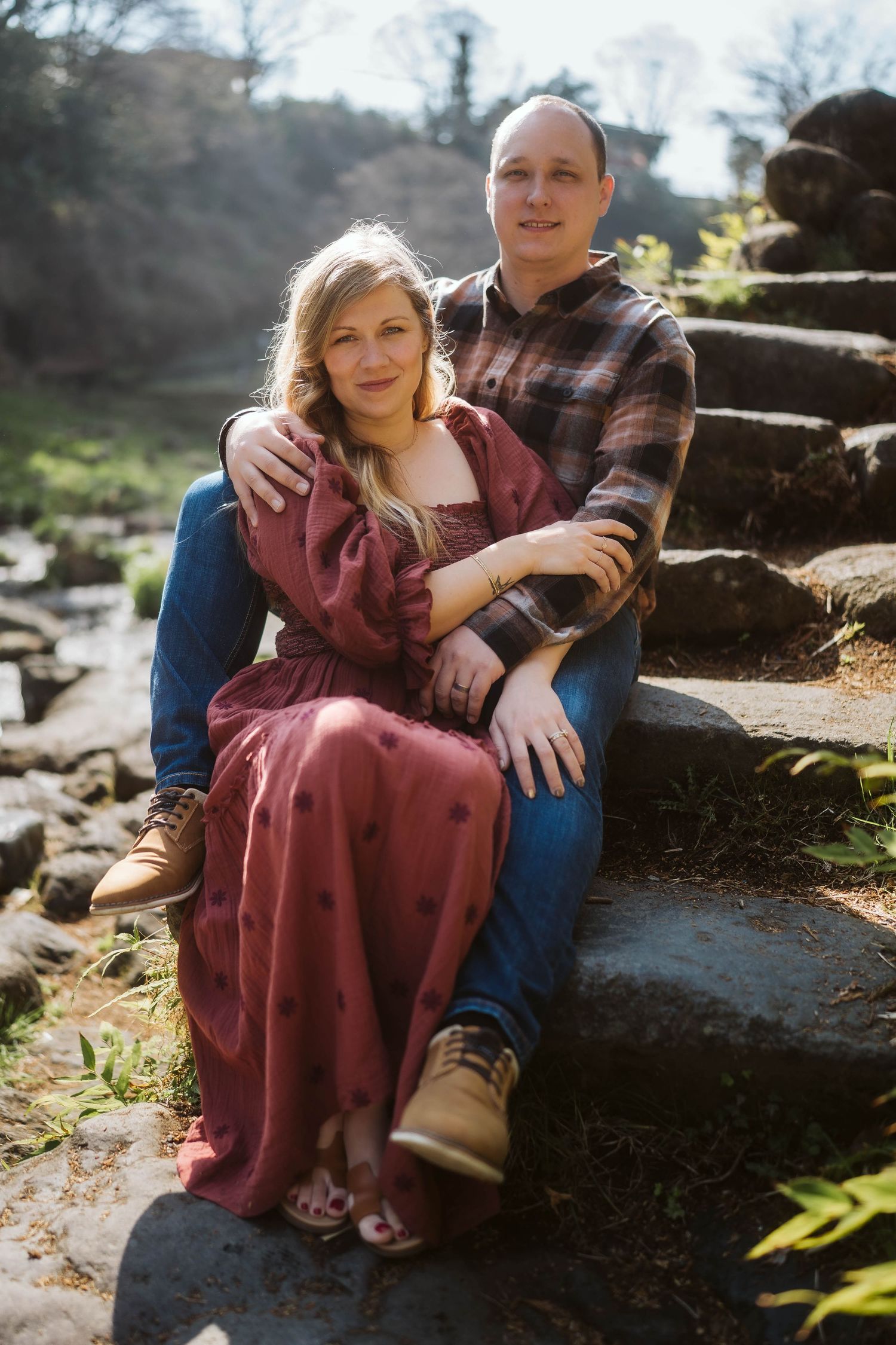 Couple sitting together on stone steps in nature wearing burgundy dress and denim during an outdoor photo session.