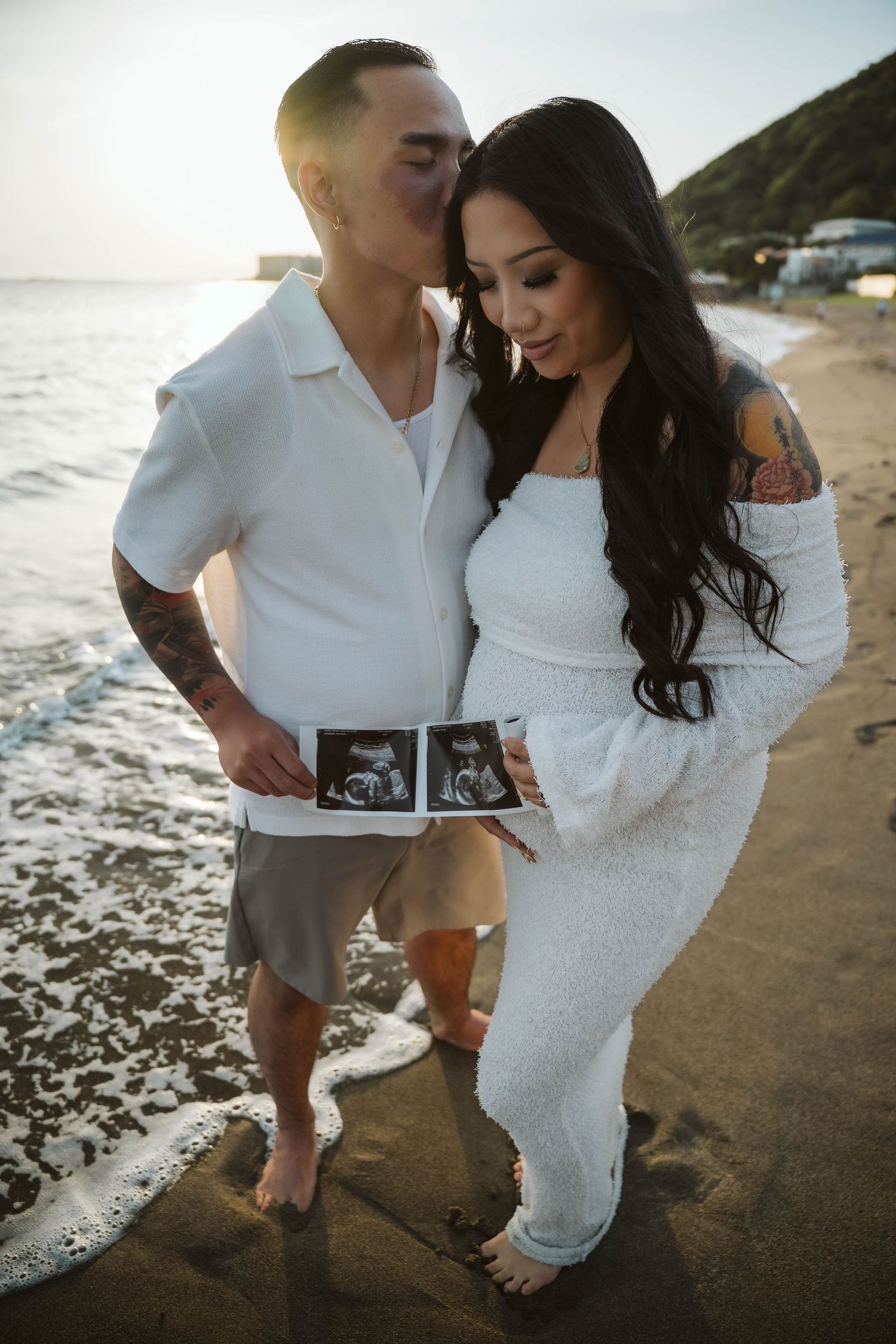 Couple shares intimate moment on beach while holding ultrasound photos during pregnancy announcement shoot.