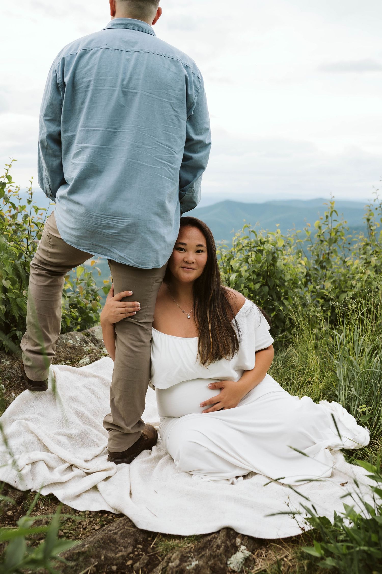 A couple shares a tender moment on a grassy mountainside, with rolling hills visible in the misty background.