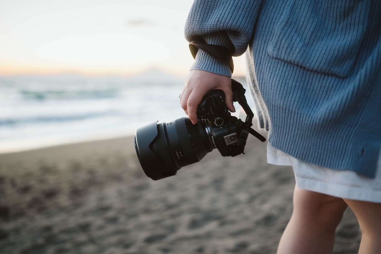 Close-up shot of hands holding a professional DSLR camera with telephoto lens on a sandy beach at sunset.