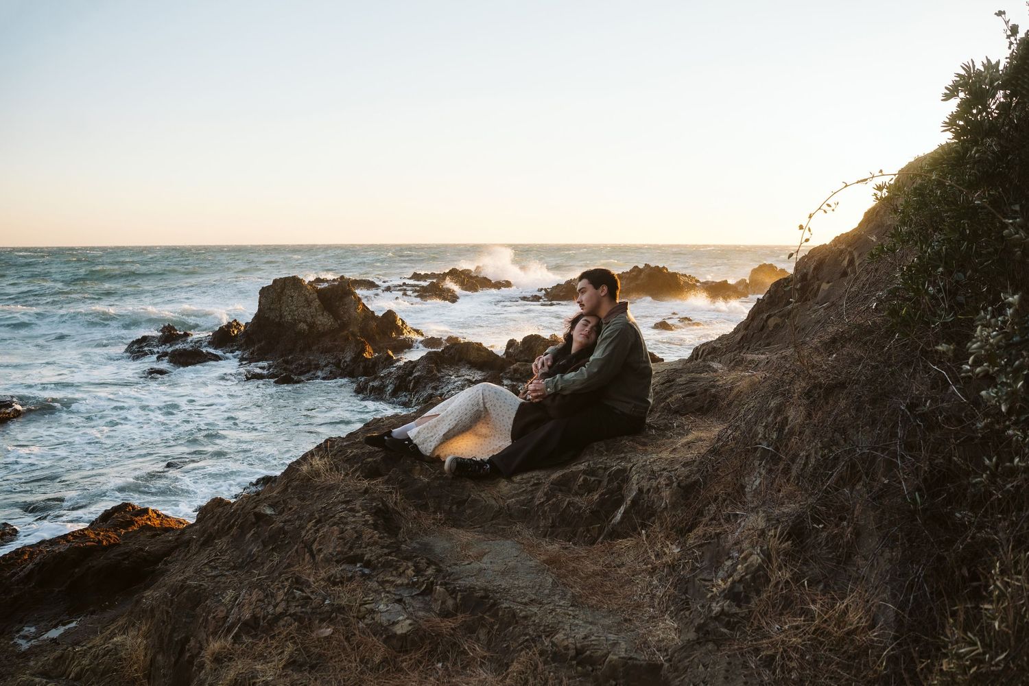 A seagull perches on rocky coastal cliffs at sunset with crashing waves in the background.
