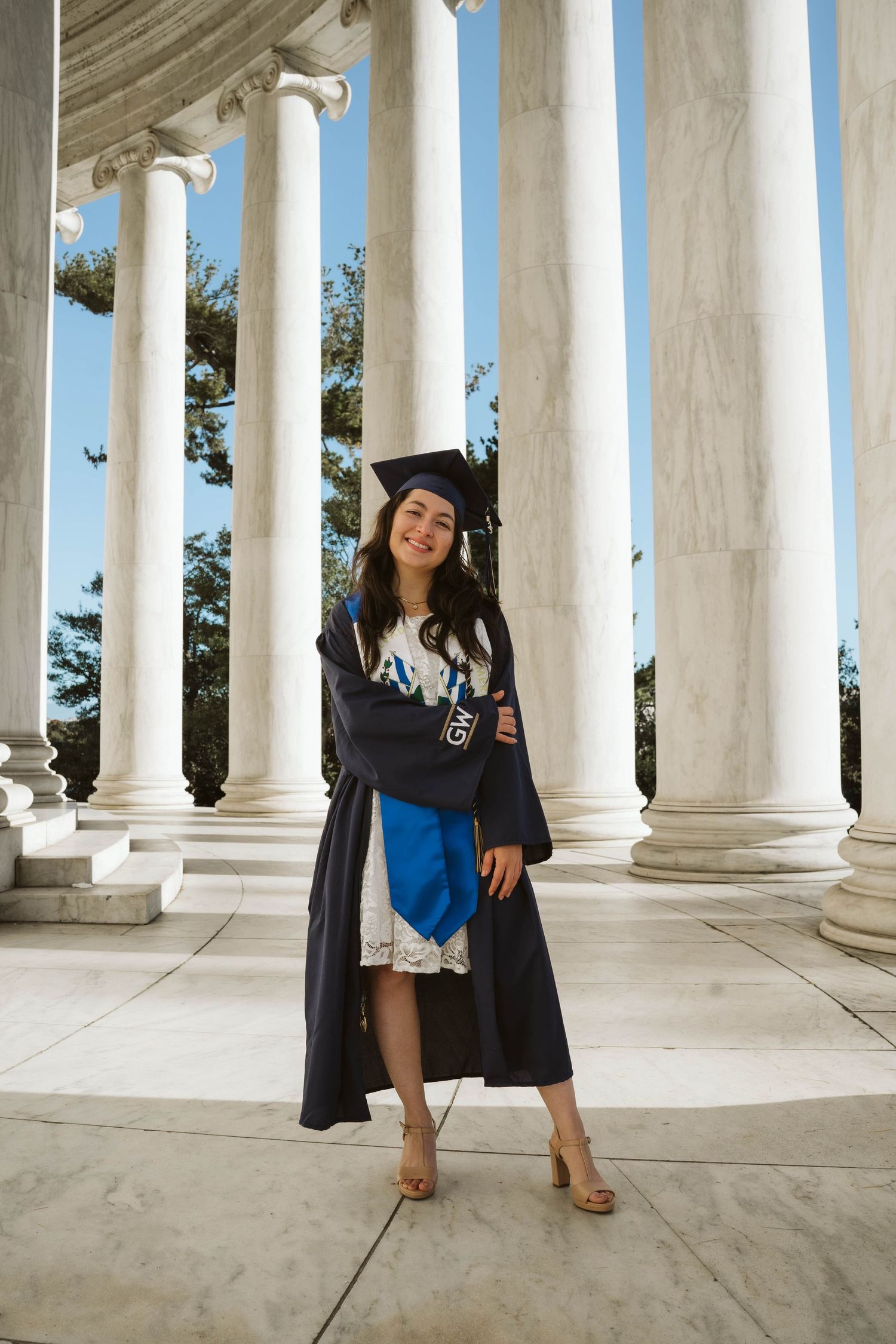 Graduate in cap and gown poses between classic white columns during outdoor graduation photo session at university.