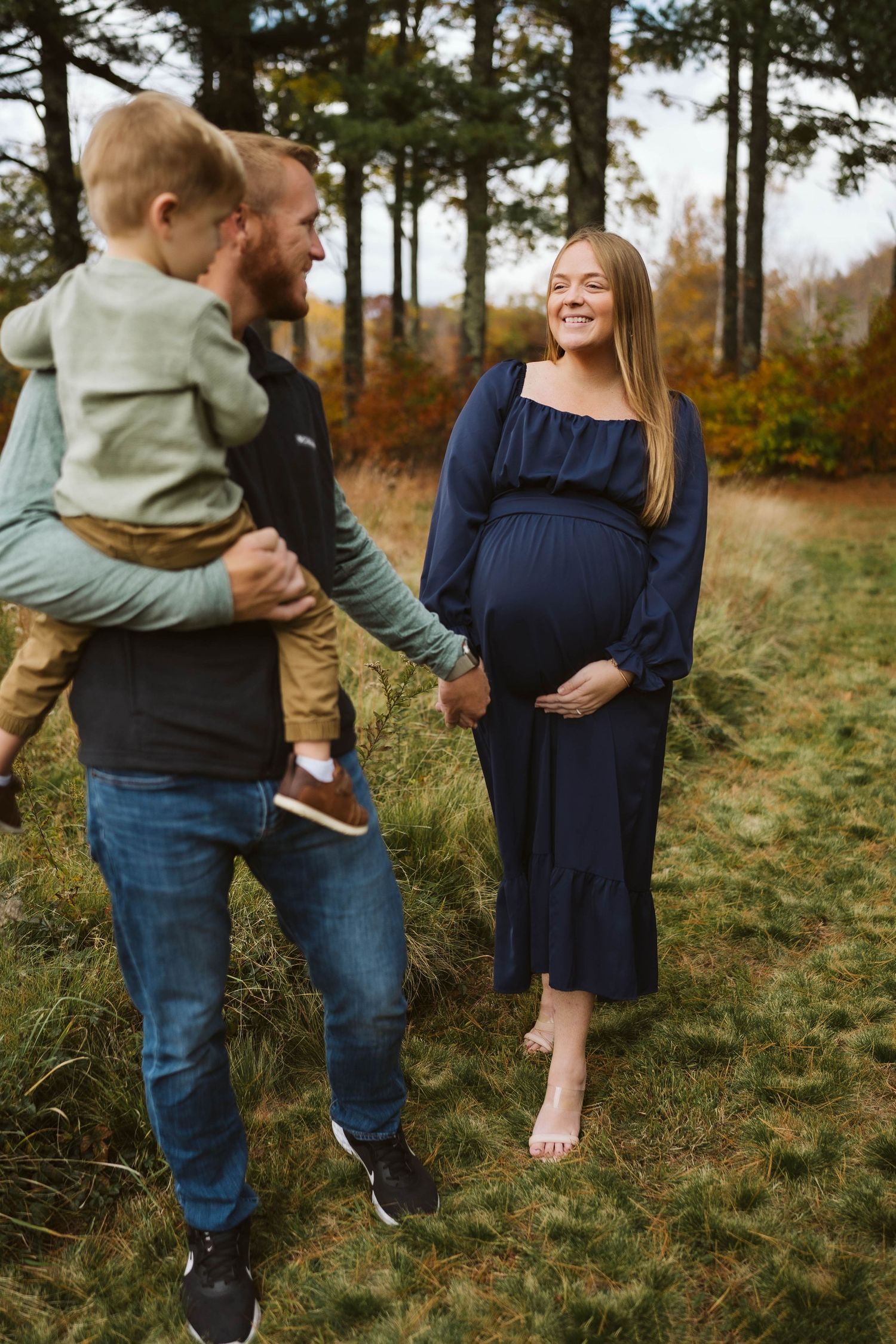 A tender family moment captured outdoors with pine trees and fall foliage in the background.