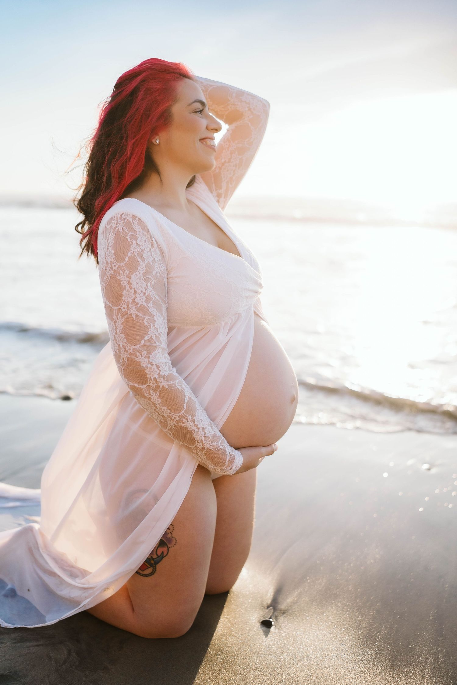 Glowing sunset maternity photoshoot on a beach with white lace dress and soft natural lighting.