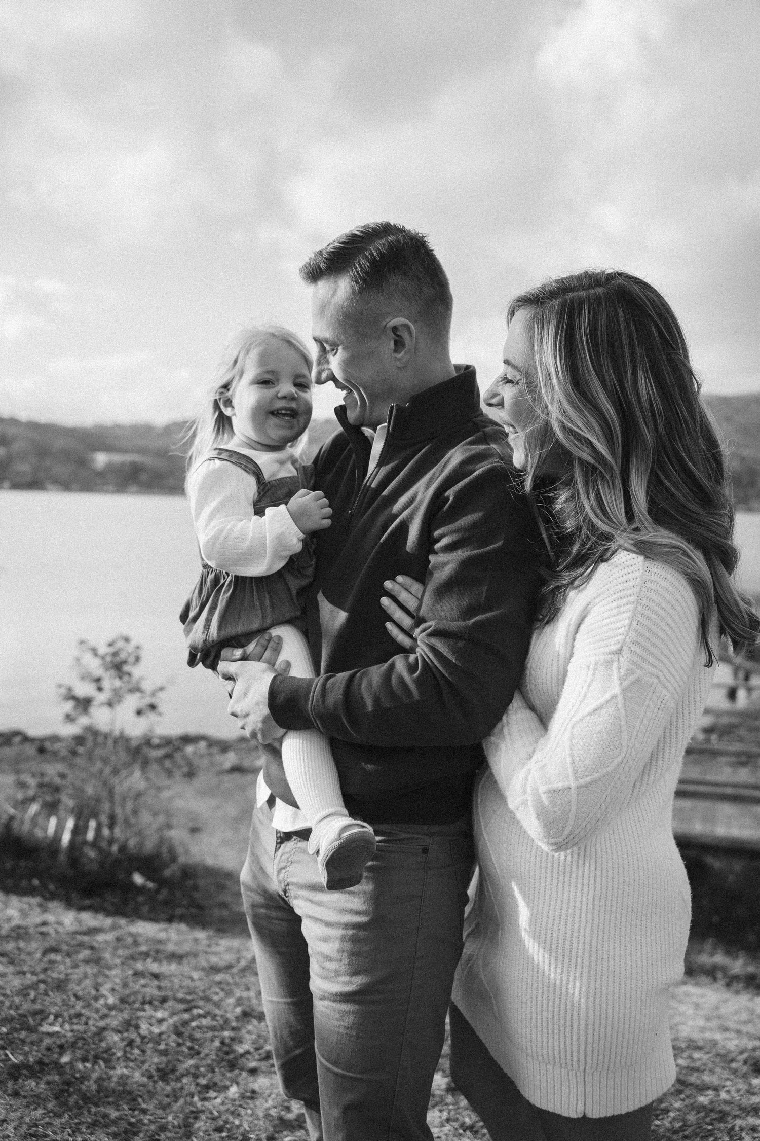 A family of three embraces outdoors in black and white photos by a scenic waterfront landscape.