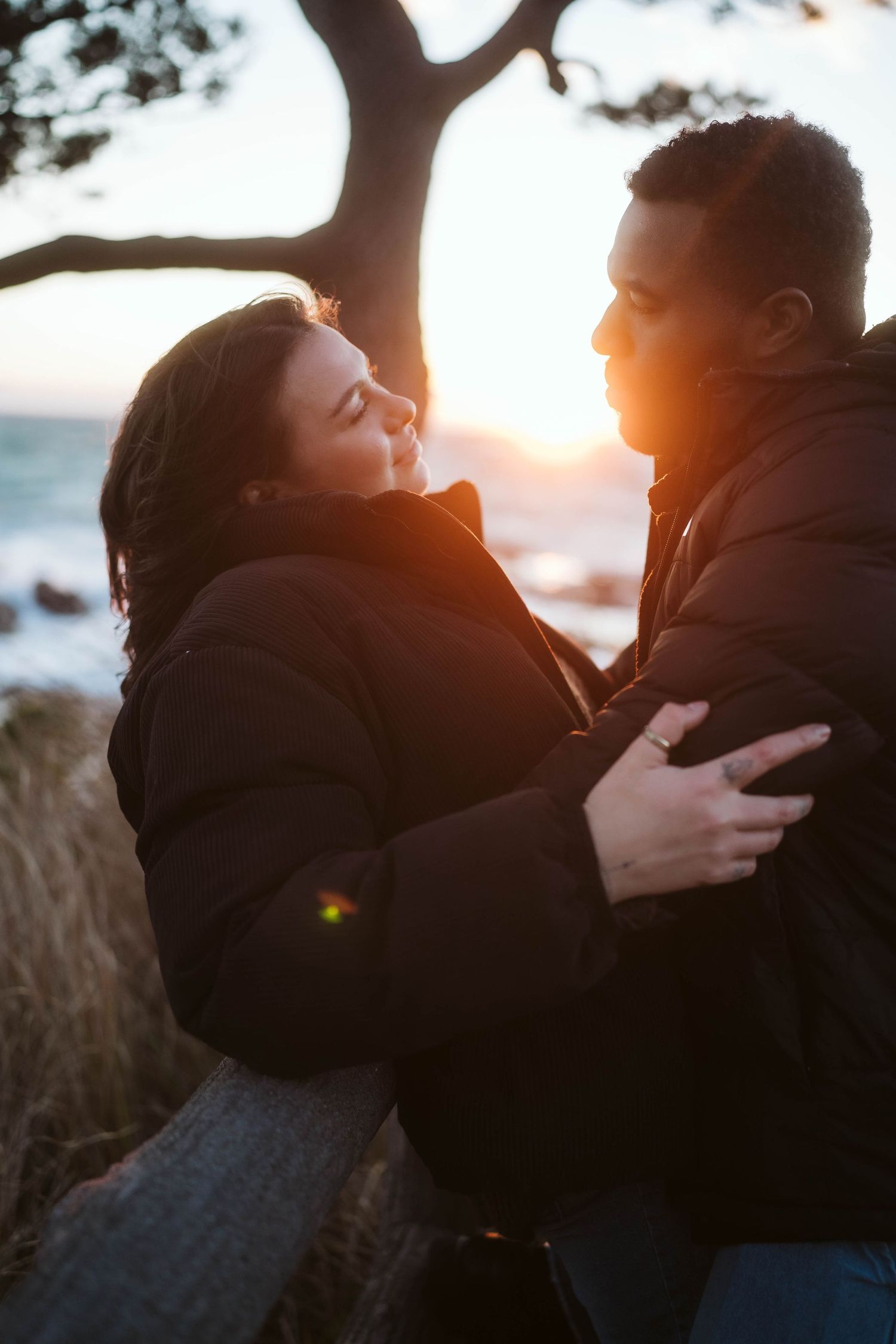 Silhouetted couple embracing outdoors at sunset with golden light streaming through trees in background.