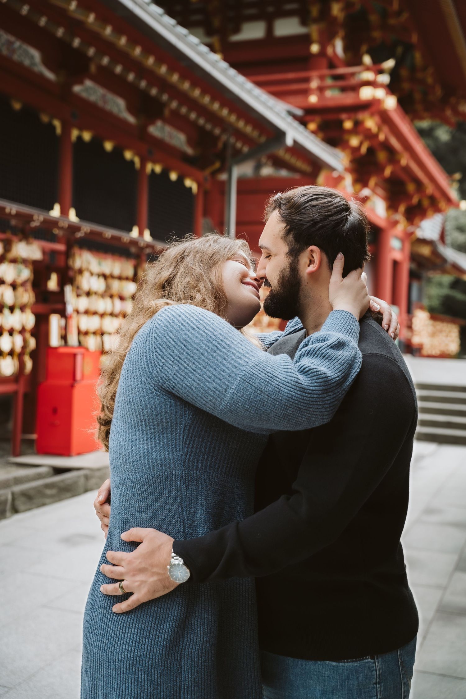 A romantic couple embraces in front of a traditional Japanese shrine with red architecture and lanterns in the background.