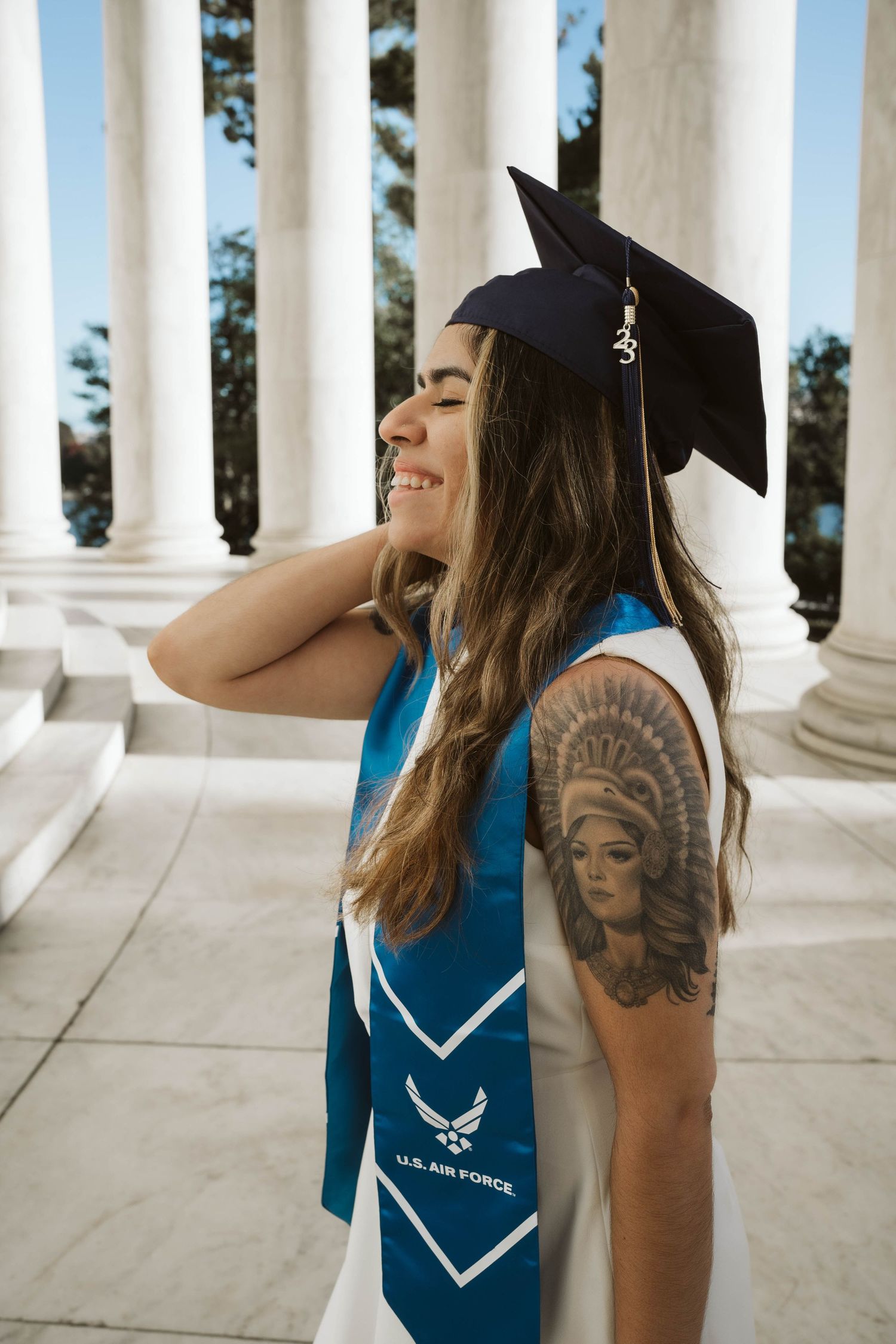 Graduate wearing Air Force stole and cap poses with tattoo by white columns during commencement celebration.