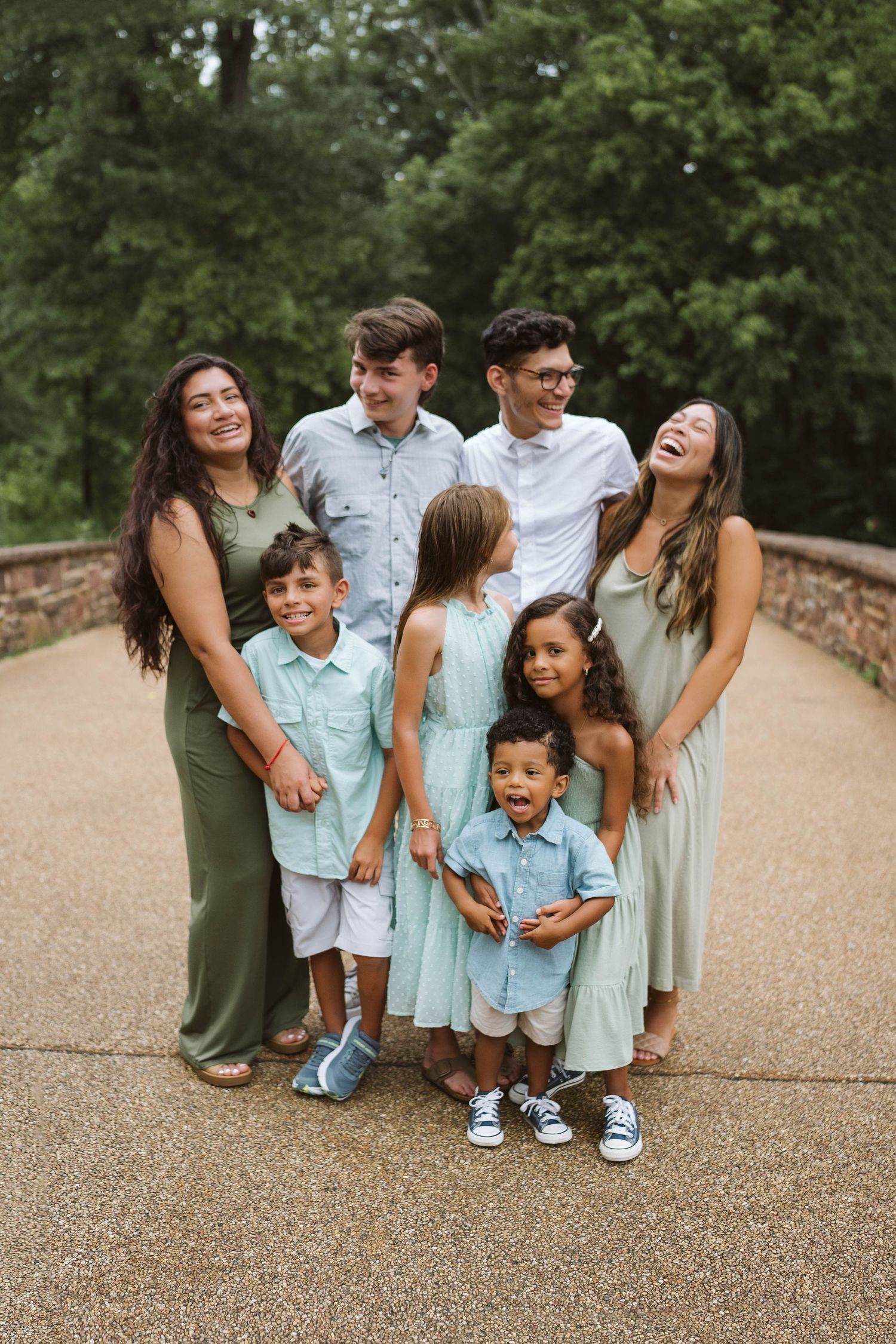 Family group wearing coordinated mint and sage outfits stands together laughing on stone path in park setting.