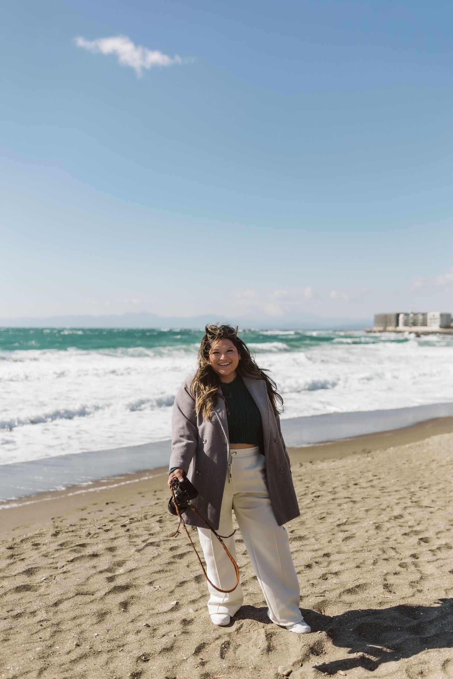 Casual beach style photoshoot featuring cozy layers and white pants against ocean waves.