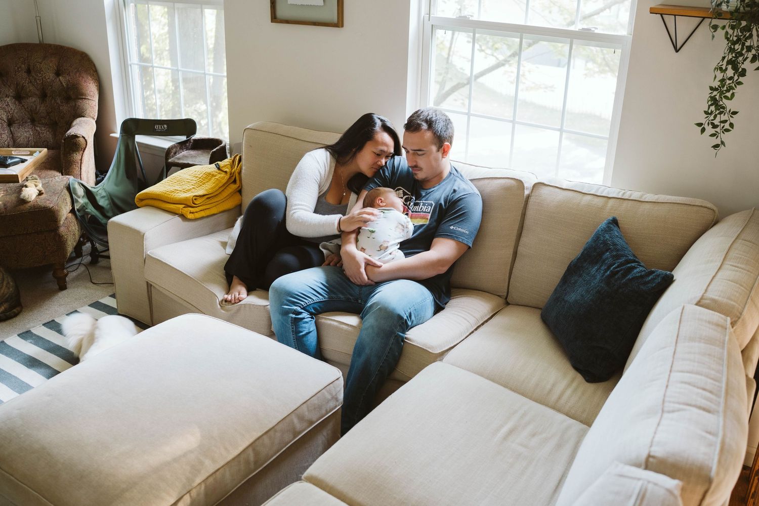 Parents cuddle with their newborn baby in a cozy living room on a beige sectional sofa near sunlit windows.