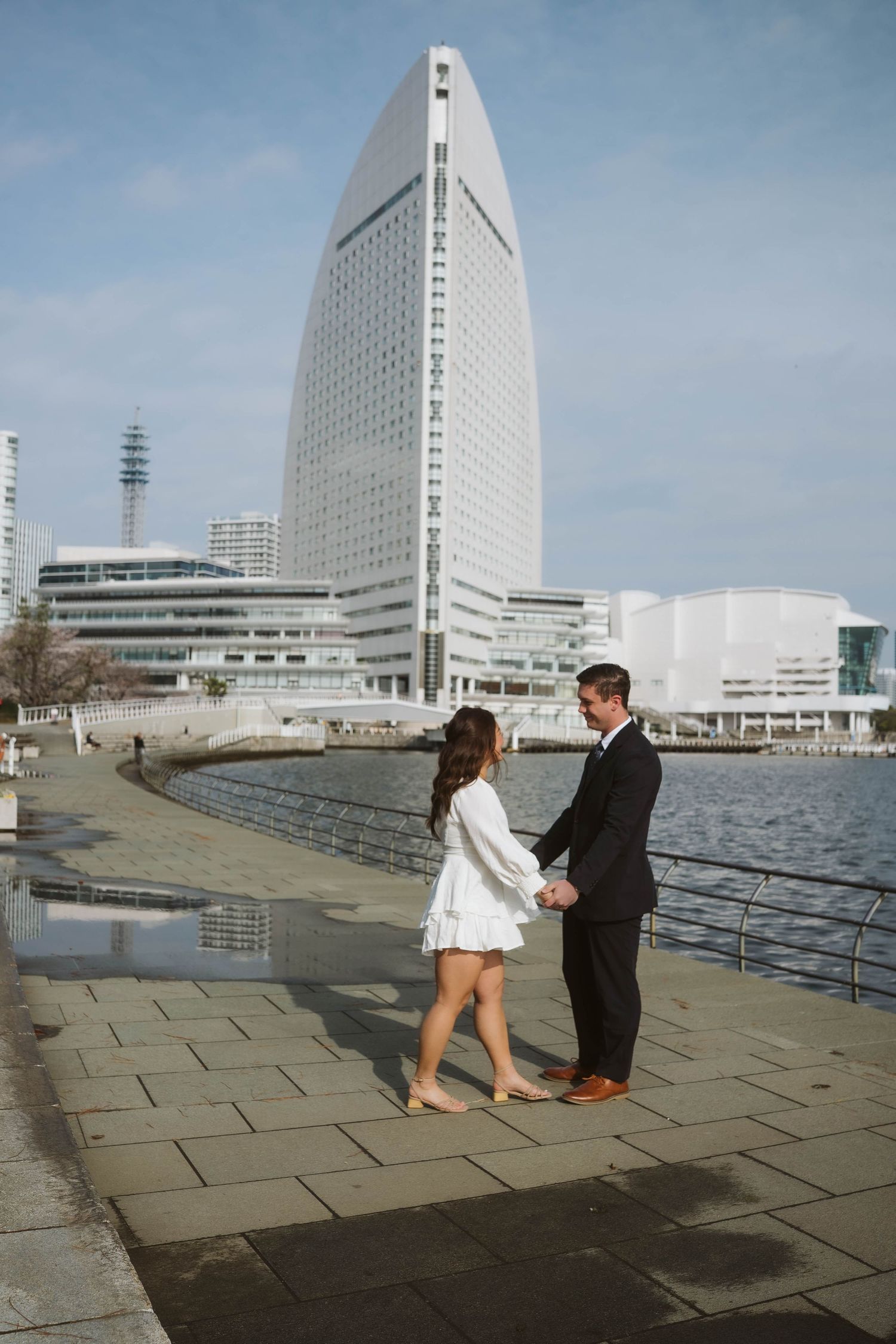 A couple holds hands by the waterfront with a modern skyscraper rising dramatically in the background.