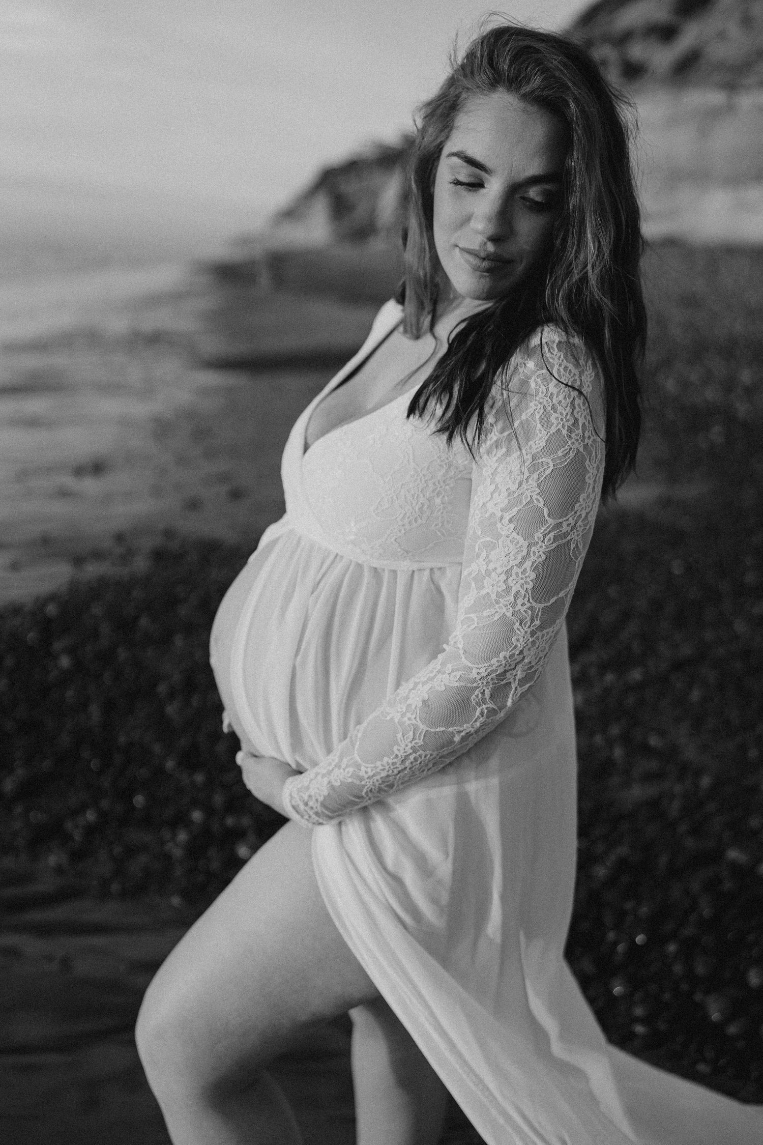 Black and white maternity photo of someone in a white lace dress posing on a rocky beach at sunset.