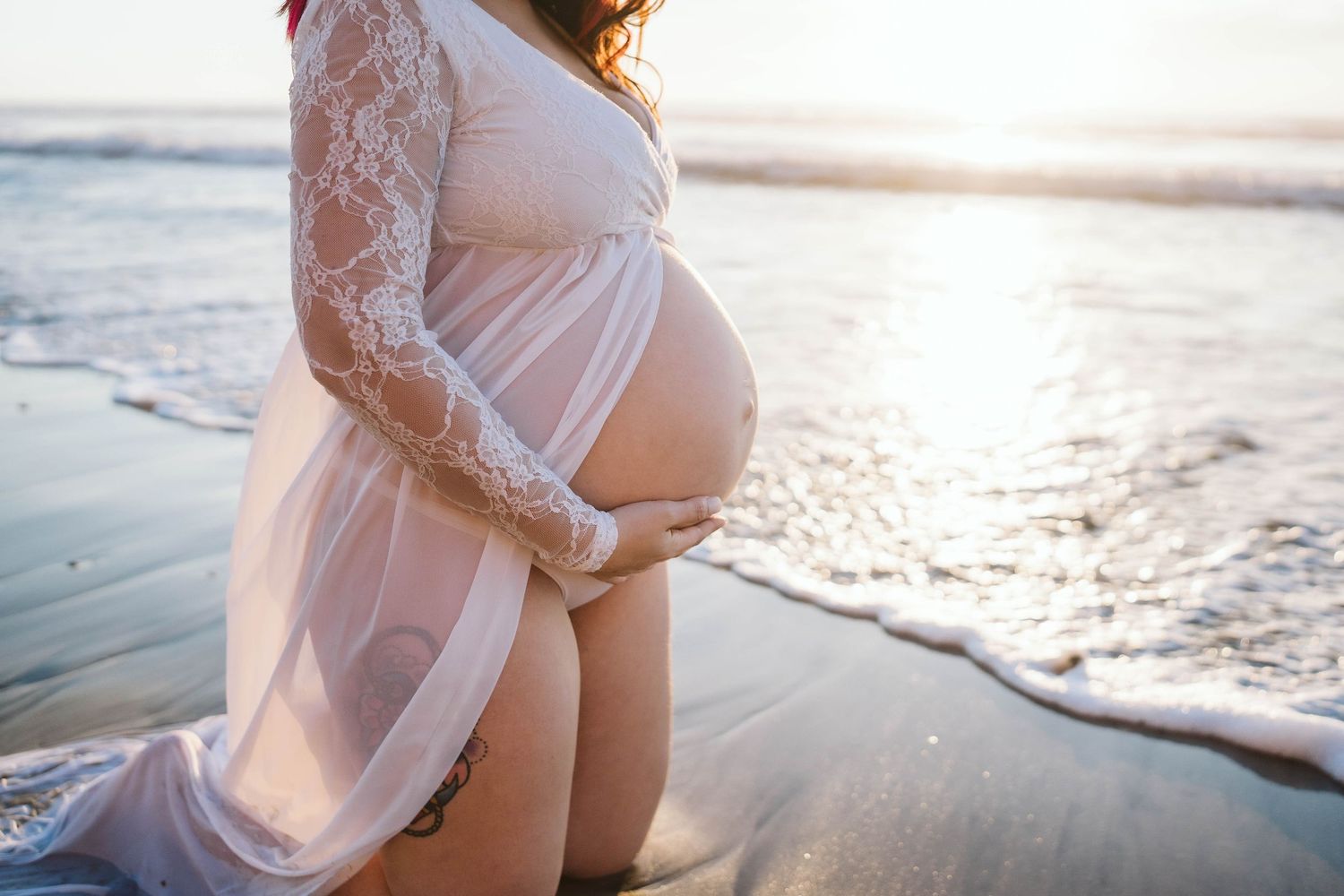 Elegant maternity photo at sunset showing white lace dress silhouetted against ocean waves on sandy beach.