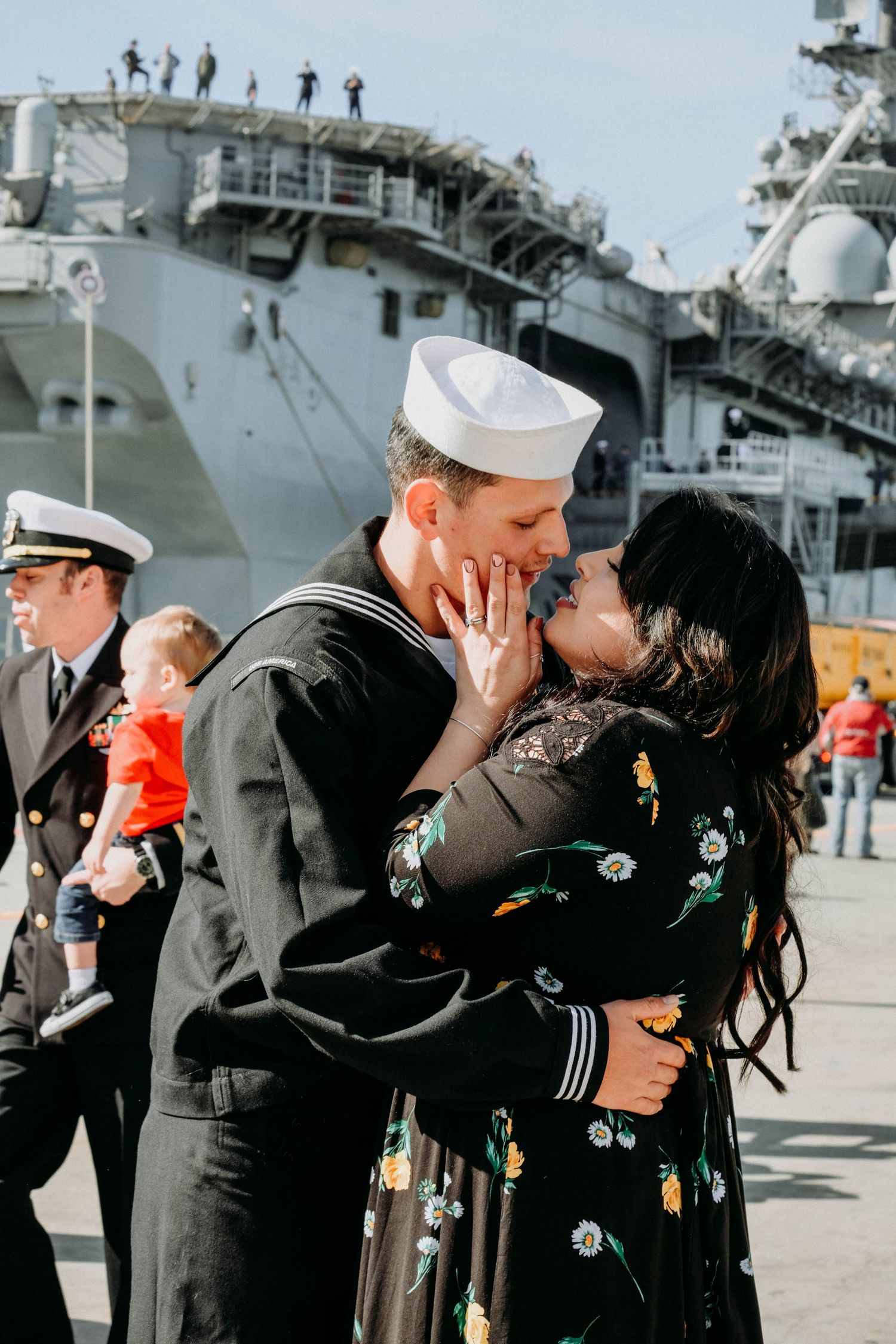 Military sailor reunites with loved one in emotional embrace on pier near naval ship during homecoming ceremony.