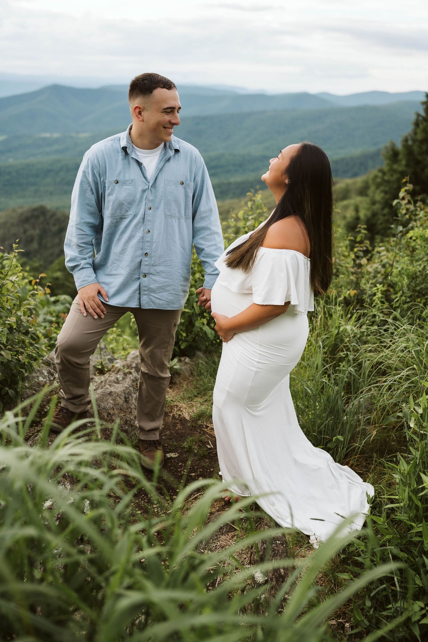 A couple stands together on a mountain overlook surrounded by lush greenery, the woman wearing a flowing white dress.