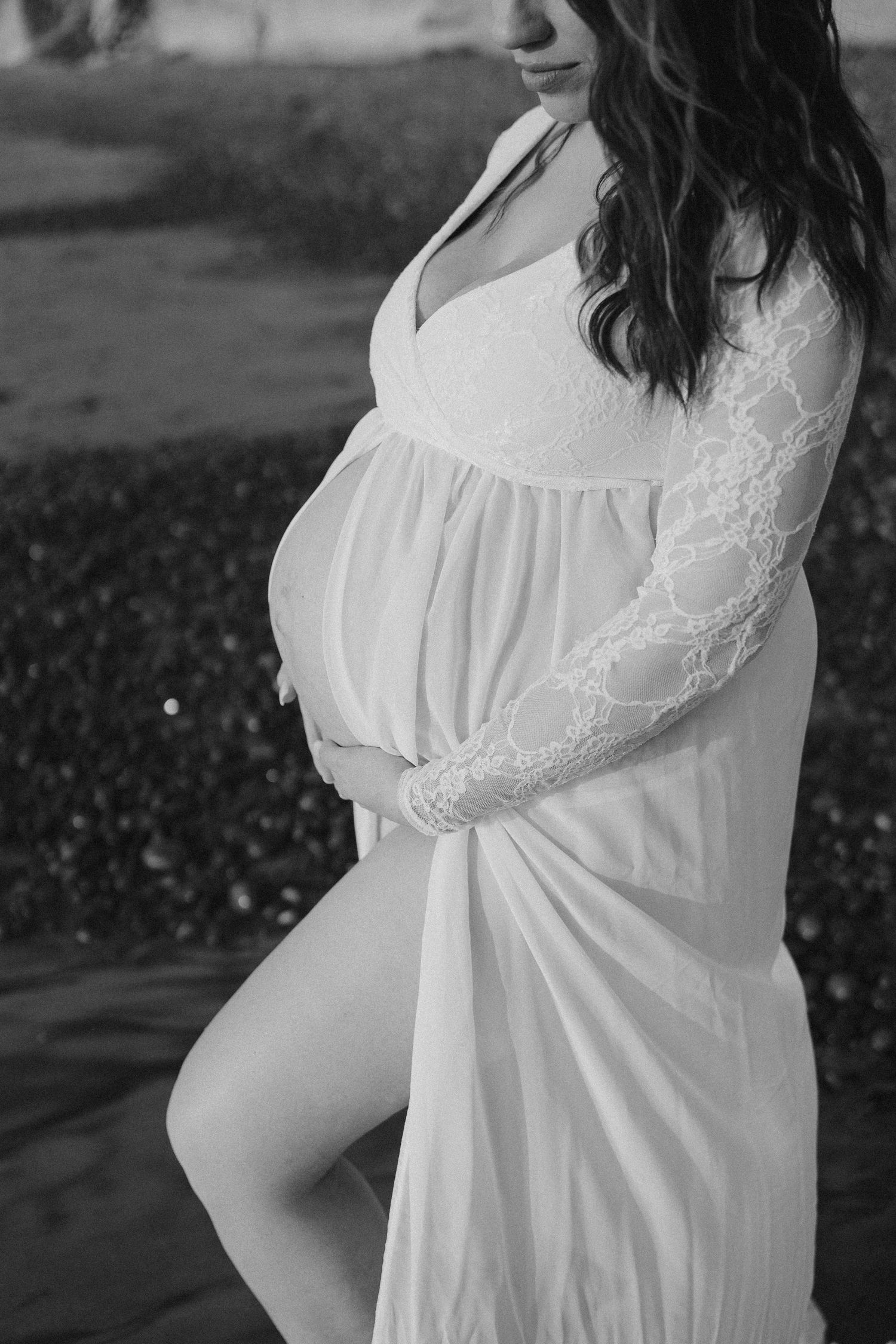 A pregnant woman wearing a white lace-trimmed maternity dress poses elegantly in a black and white photo.