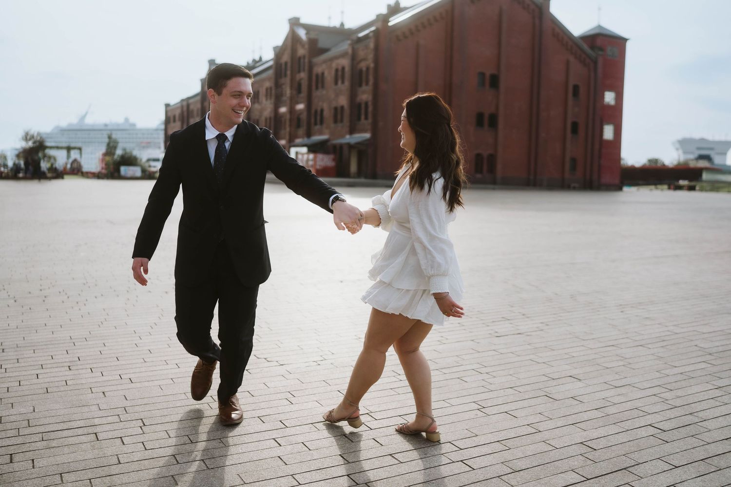 A couple in formal attire dances together on a brick plaza outside historic red brick buildings on a cloudy day.