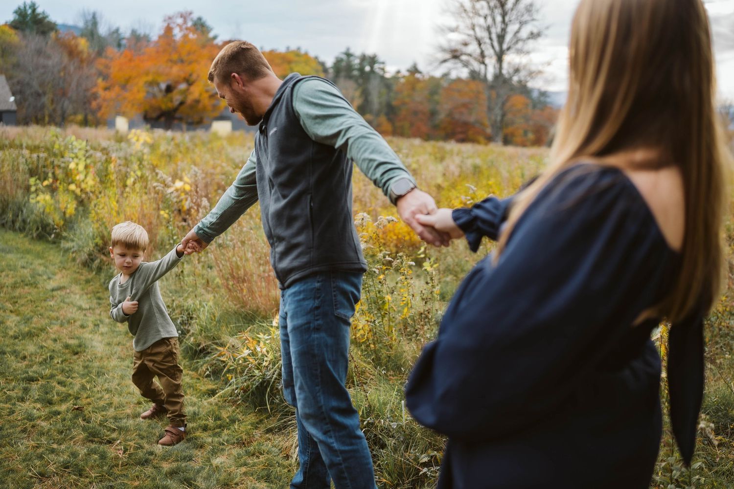 A family walks together holding hands through an autumn field surrounded by fall foliage and colorful trees.