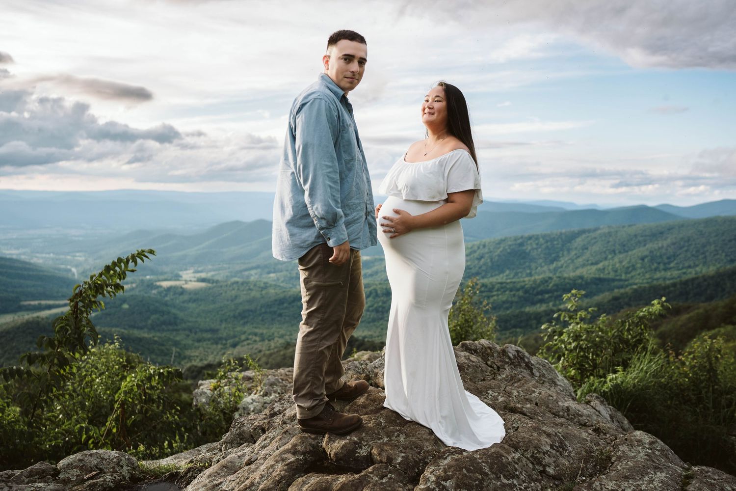 A couple stands atop a mountain overlook wearing casual and formal attire against a backdrop of rolling hills and cloudy sky.