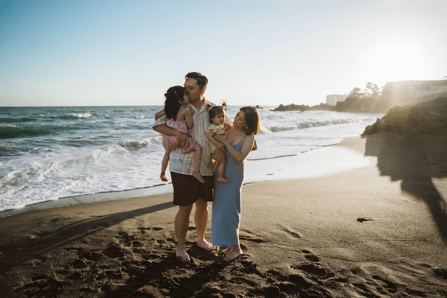 A group embraces together on a sunny beach with crashing waves and golden sunlight in the background.