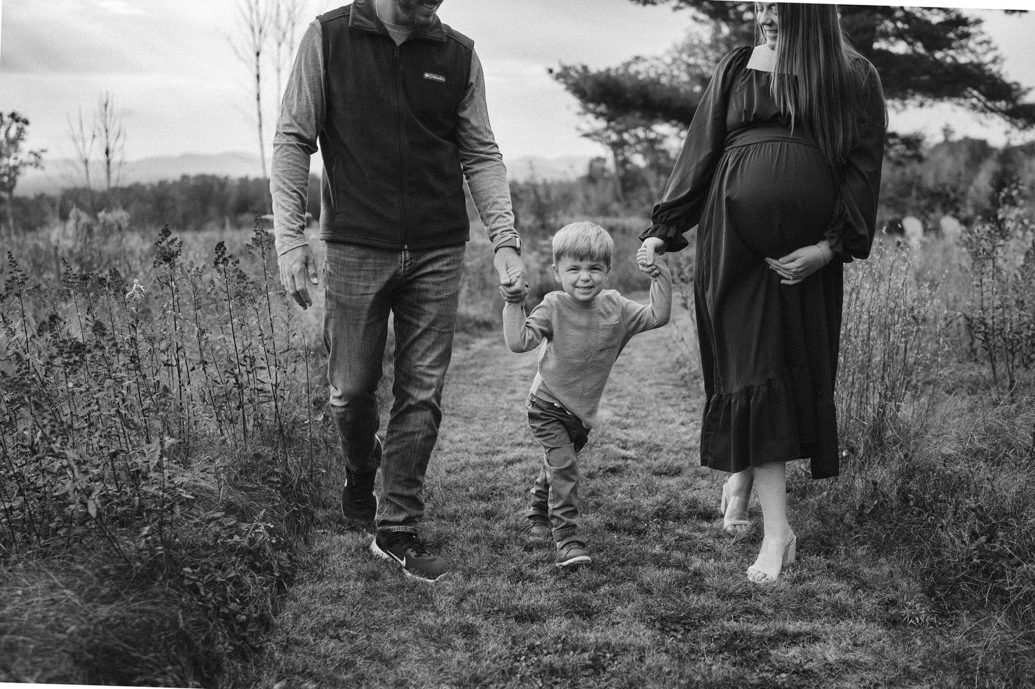 A black and white photo shows a toddler walking between two adults on a grassy rural path.