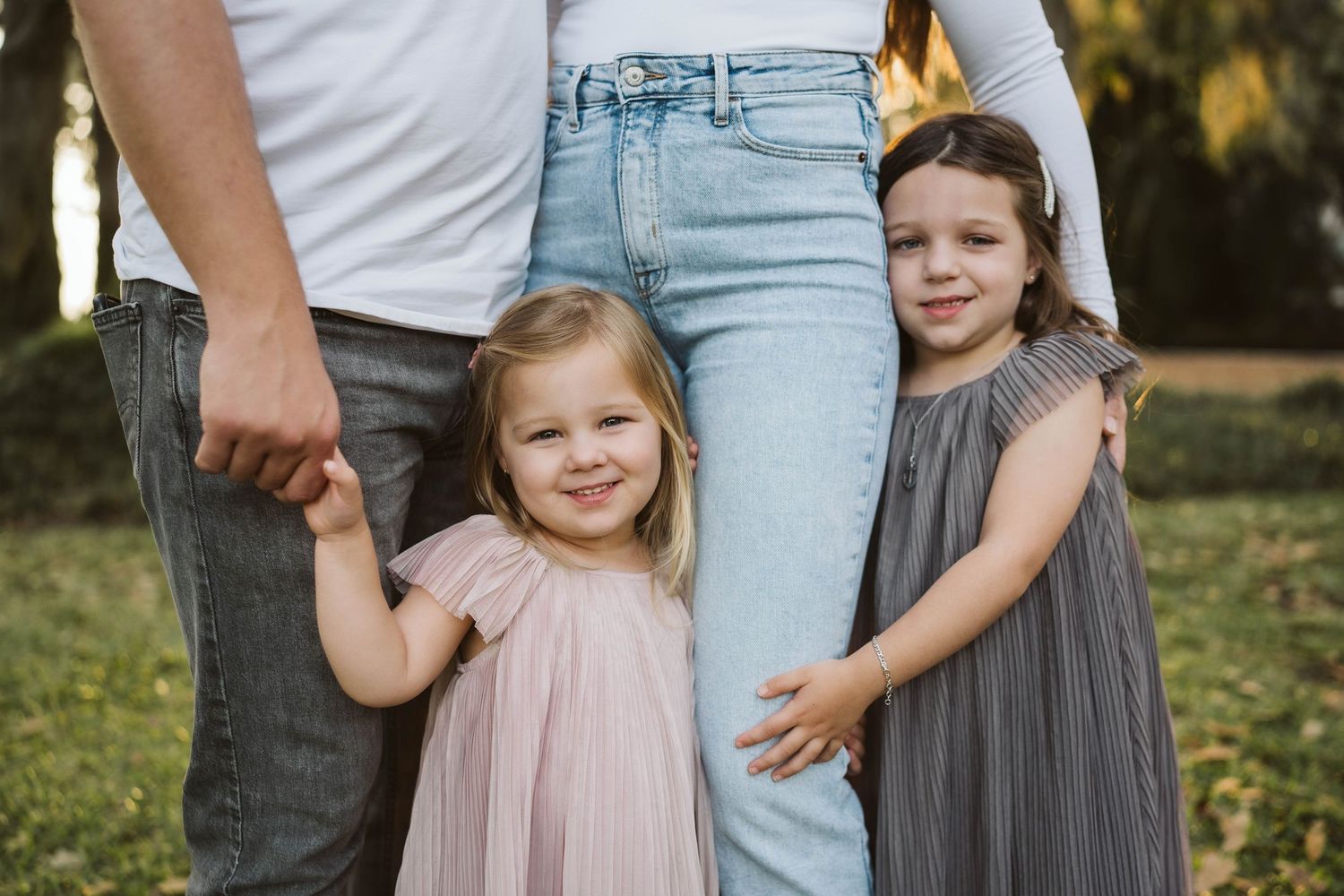 A close-up shot of children in flowy dresses holding onto their parents' legs outdoors during golden hour.