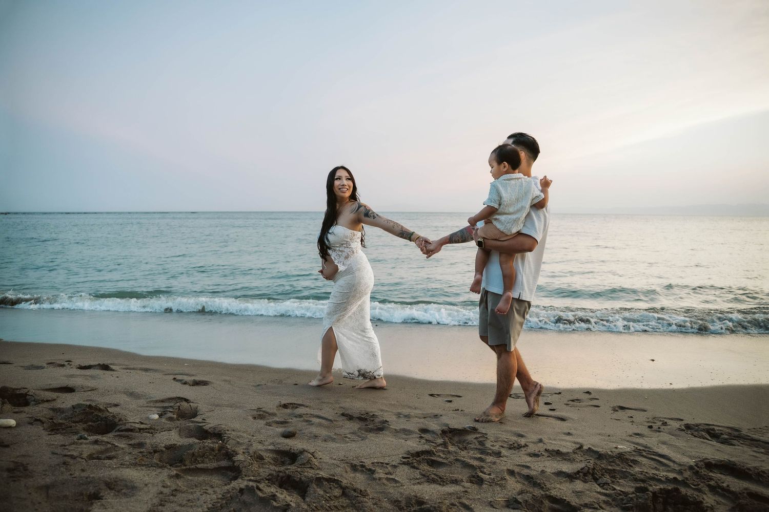 A couple in white outfits dances together on a scenic beach at sunset with ocean waves in the background.