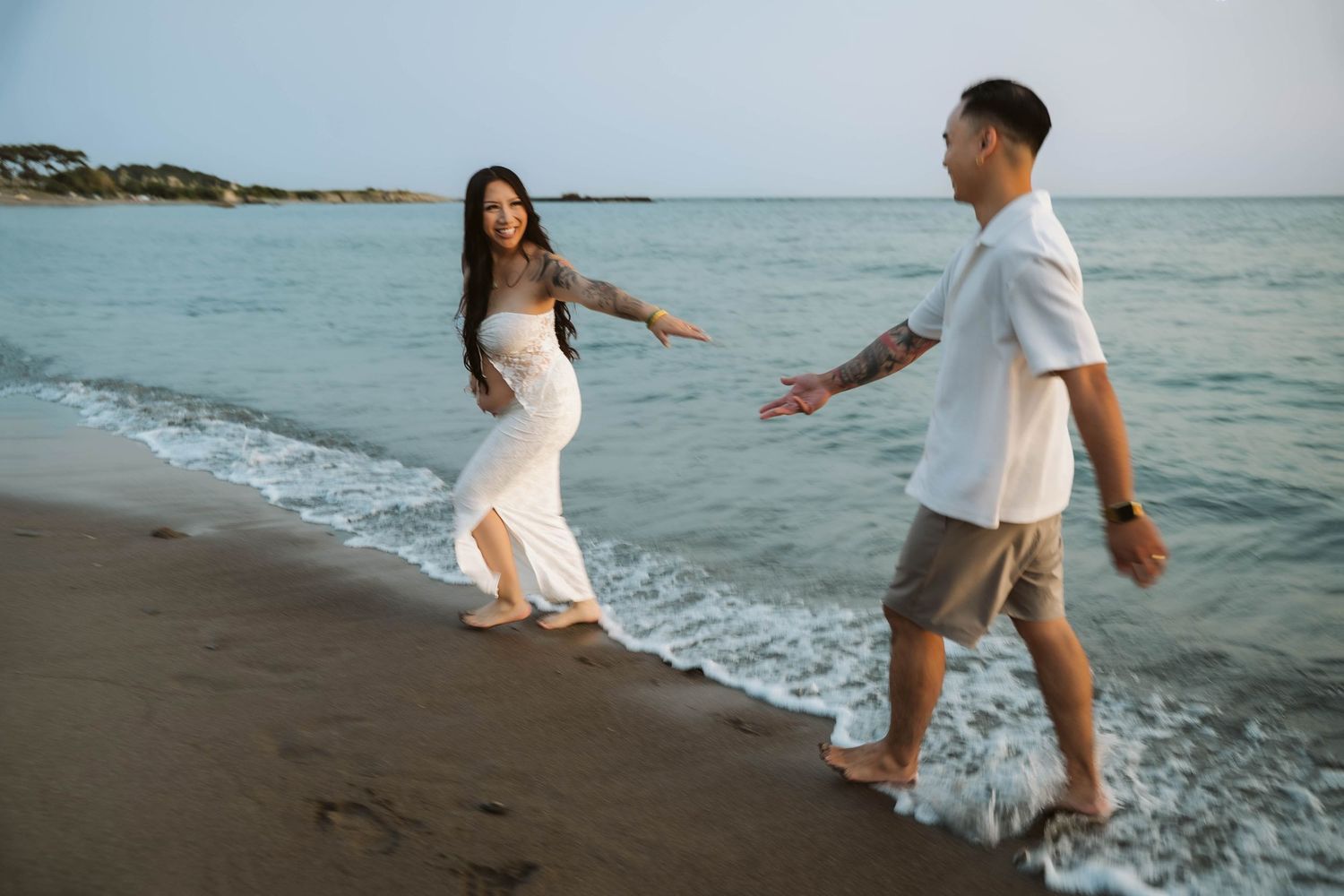 Couple in white and casual attire enjoying playful moments together on a sandy beach by the ocean.