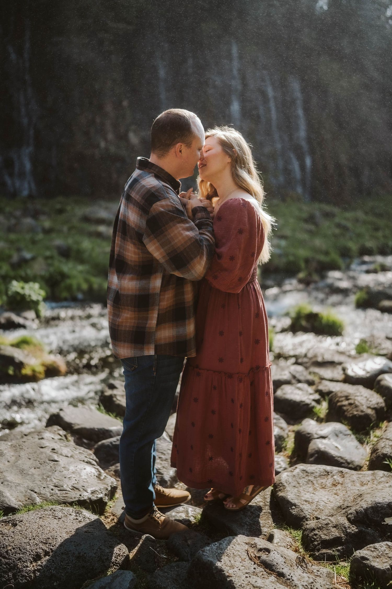A romantic couple shares a tender kiss while sitting on mossy rocks near a waterfall in a misty forest setting.