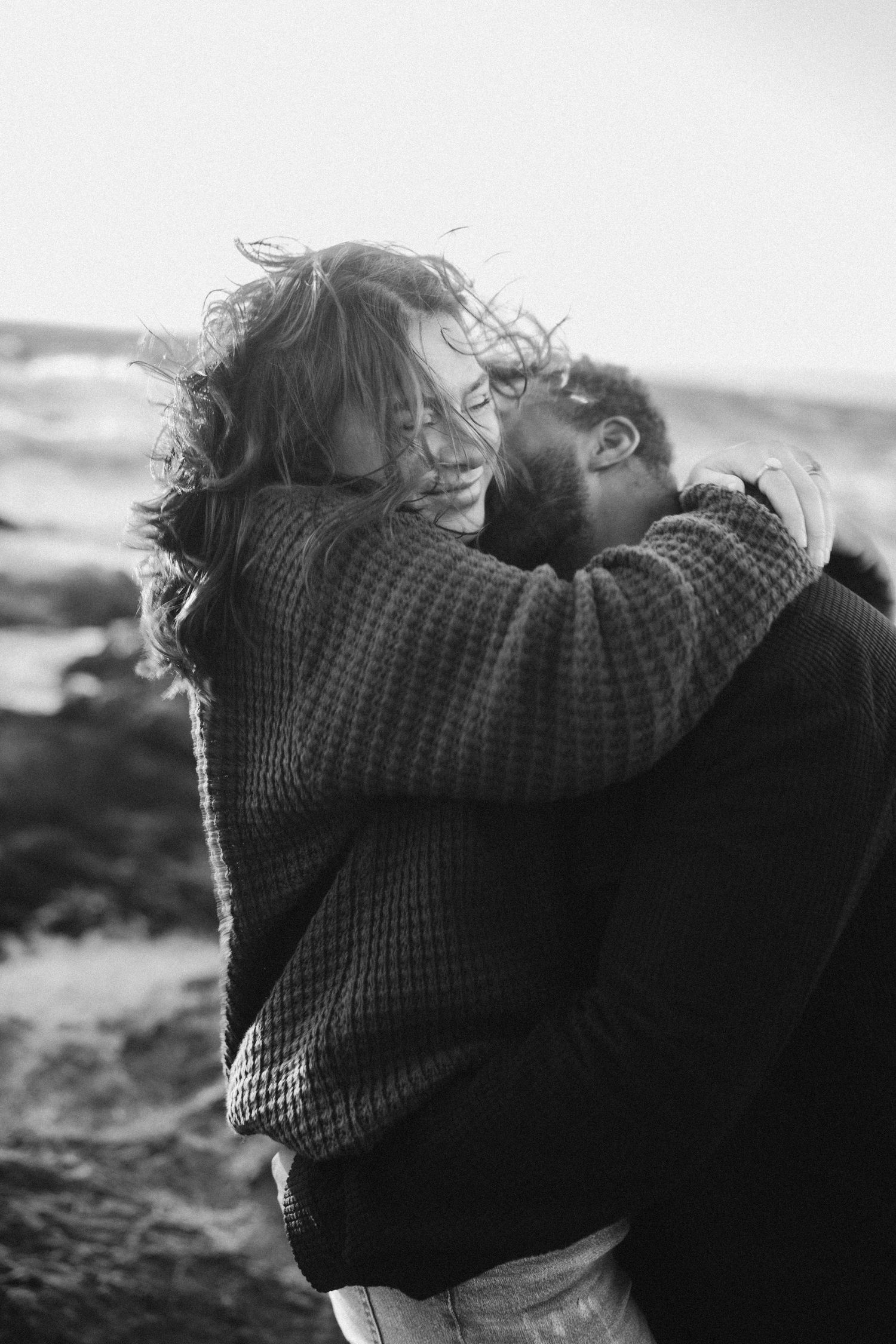 A couple embraces on a windswept coastal landscape in black and white.