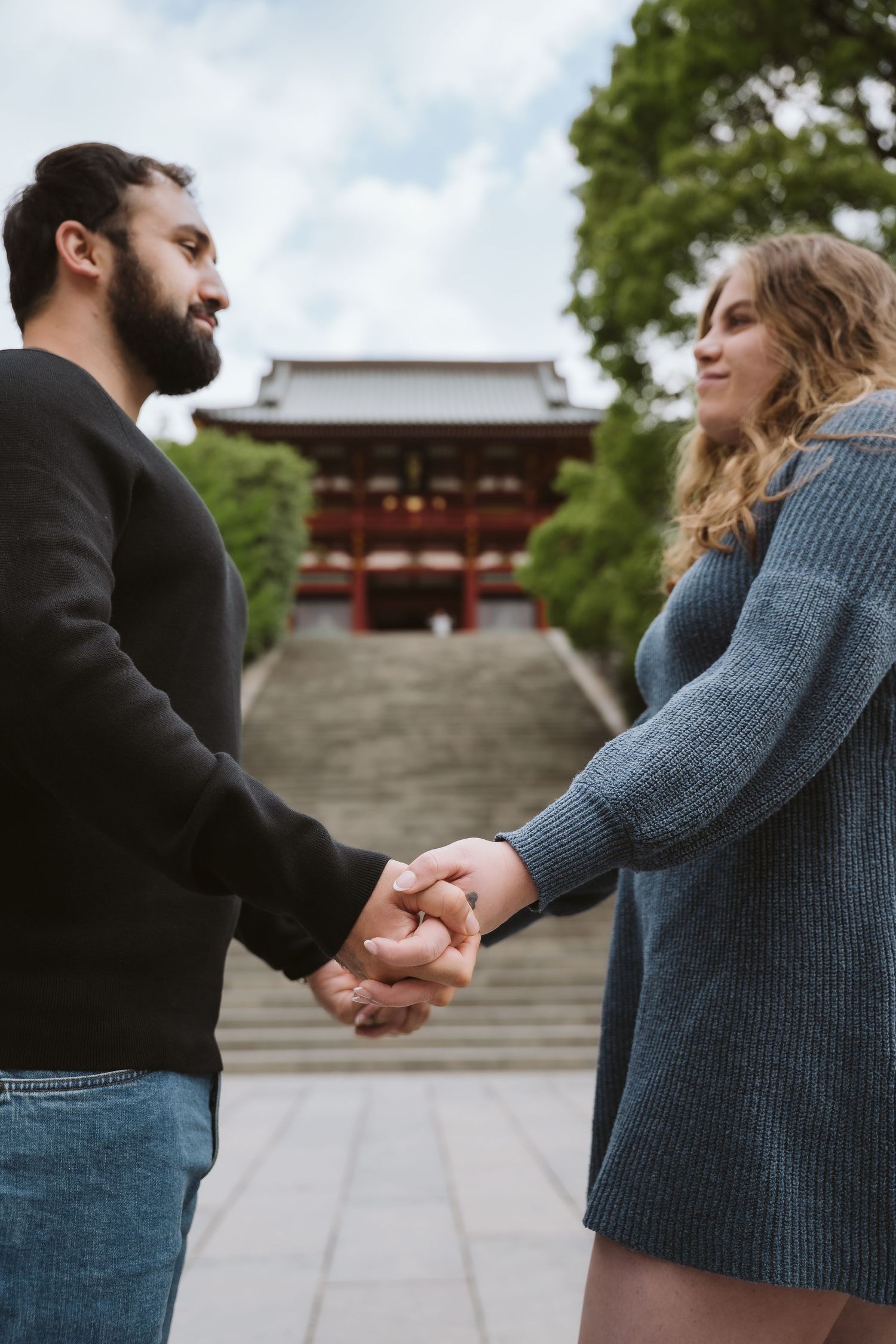 A couple holds hands while standing in front of a traditional Japanese temple gate on a stone path.