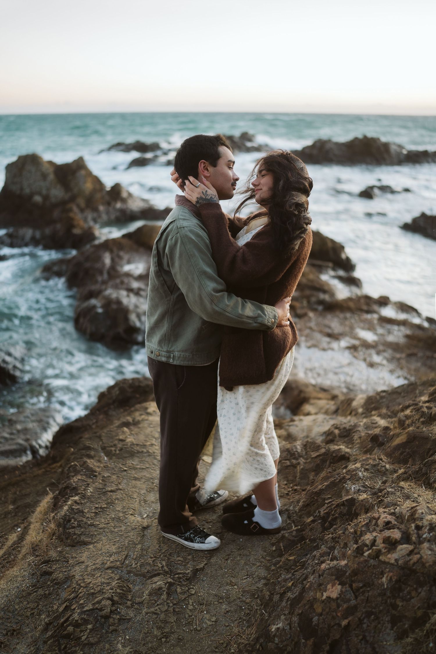 A romantic couple embraces on rugged oceanside rocks while waves crash against the shore during golden hour.