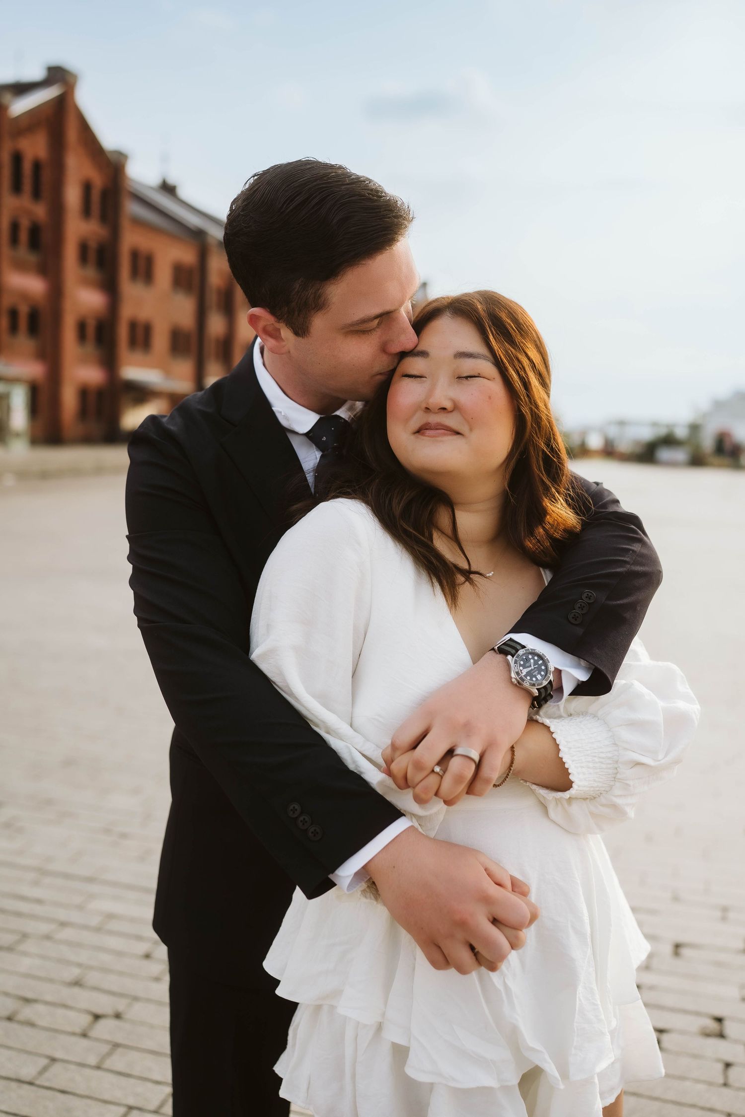 A romantic couple embraces outdoors with the bride in white and groom in black against a brick building backdrop.
