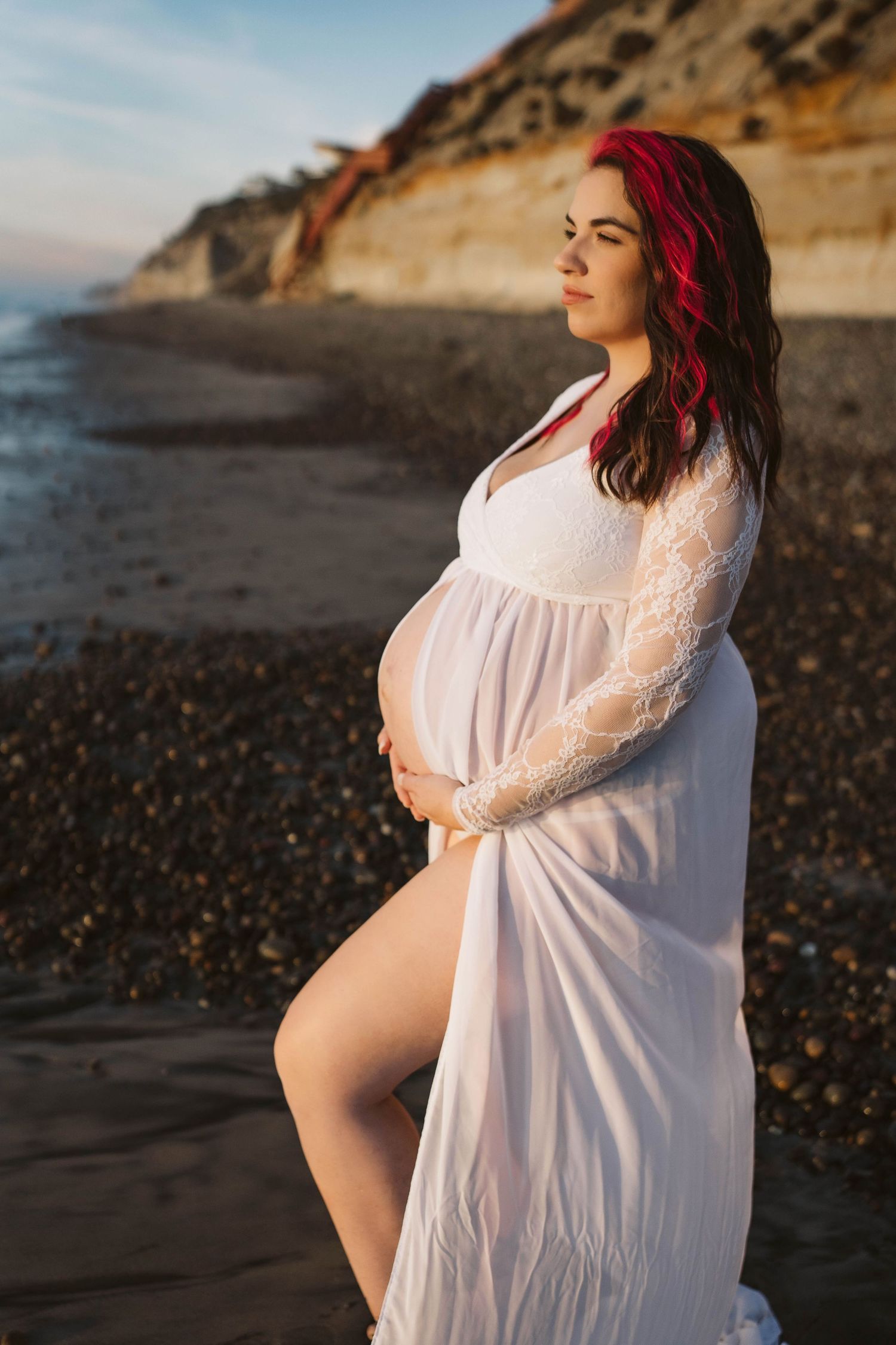 Expectant mother in flowing white dress poses on rocky beach at sunset with cliffs in background.