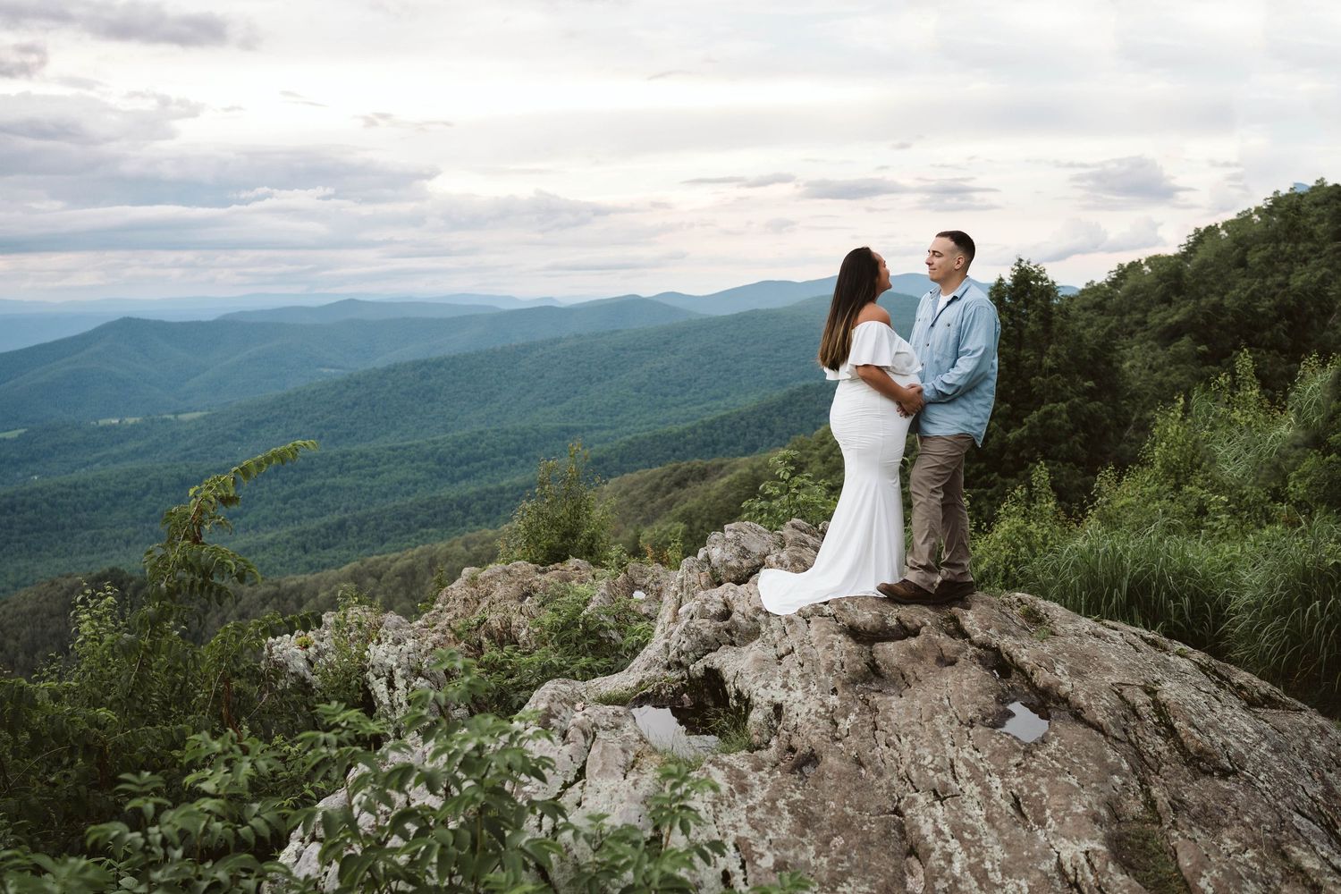 A couple in wedding attire stands together on a rocky mountain overlook with sweeping views of green rolling hills.