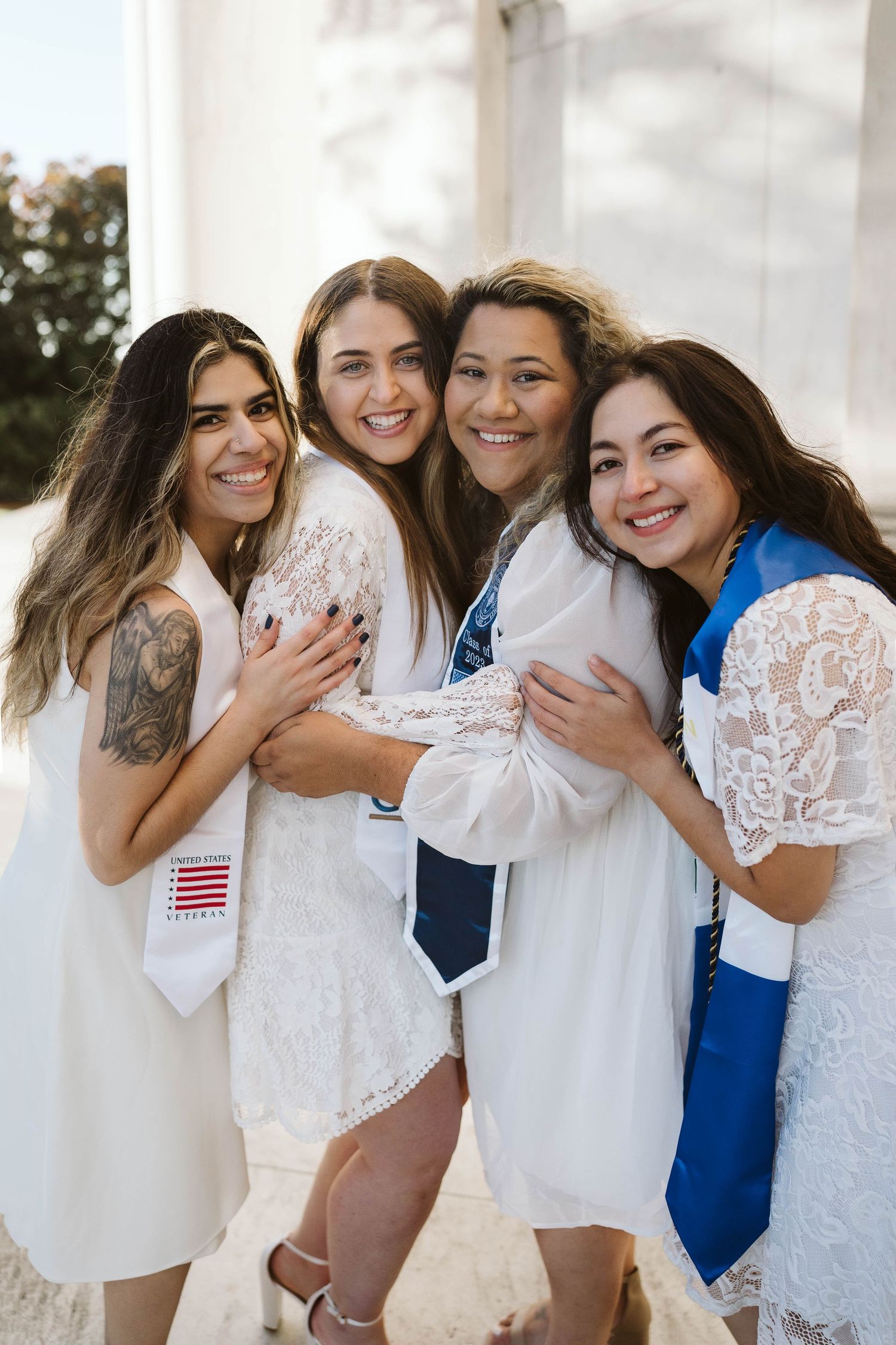 A group wearing white dresses and sashes stand together smiling and embracing in a joyful celebration.