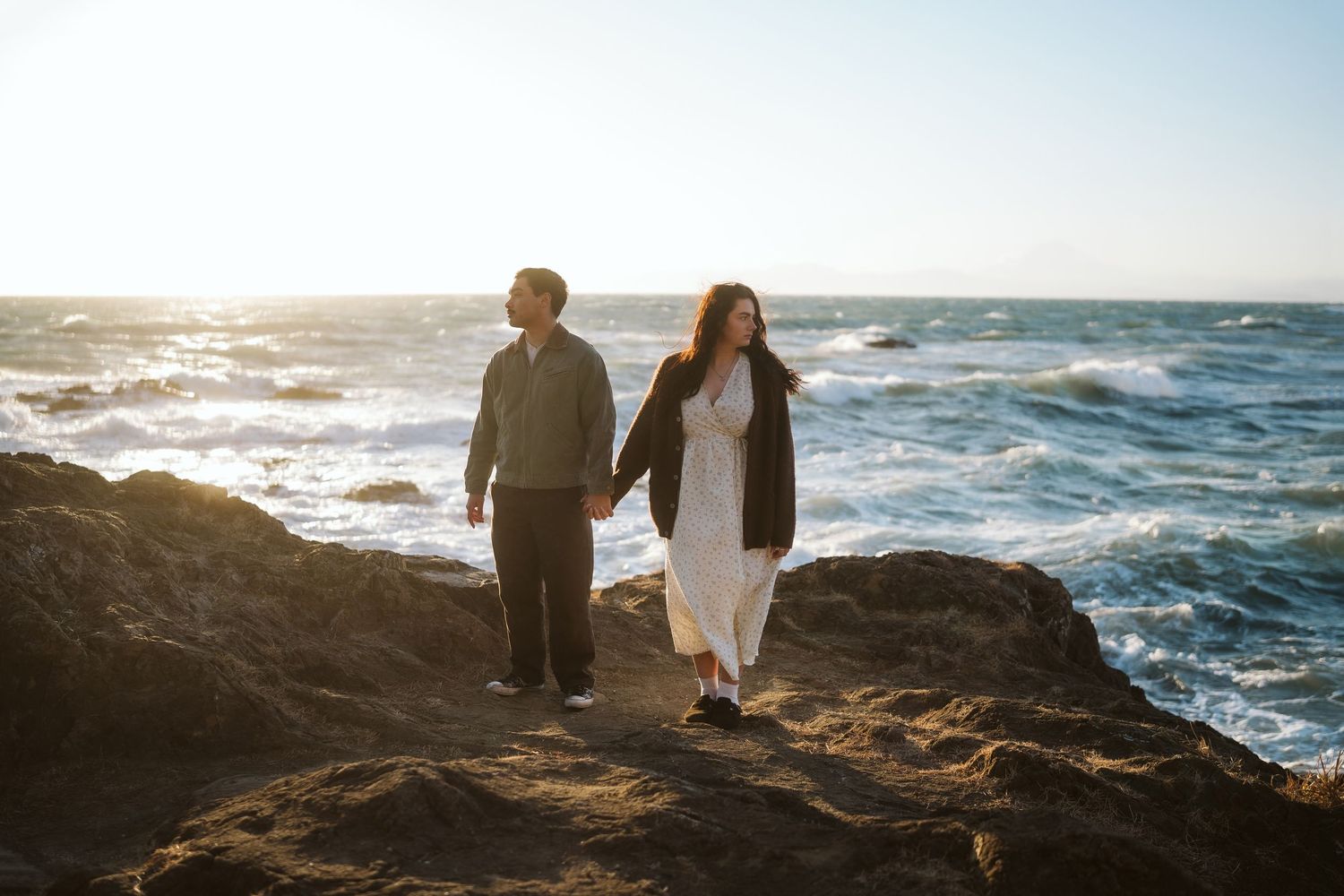 A couple stands on rocky coastal cliffs at sunset while waves crash against the shore in the background.