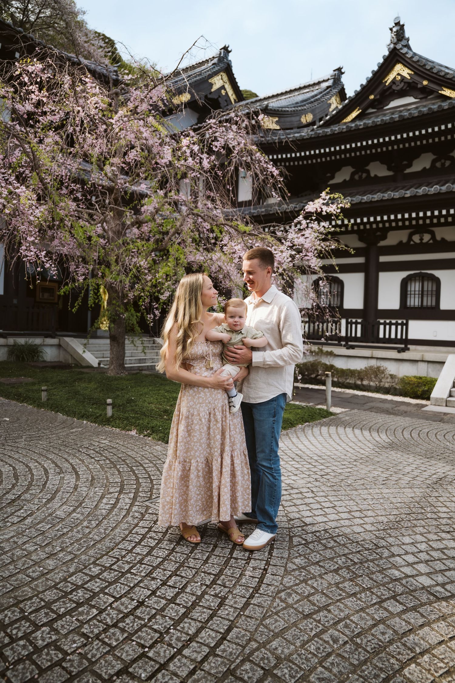A couple stands together on a circular stone path in front of a traditional Japanese temple with blooming cherry blossoms.