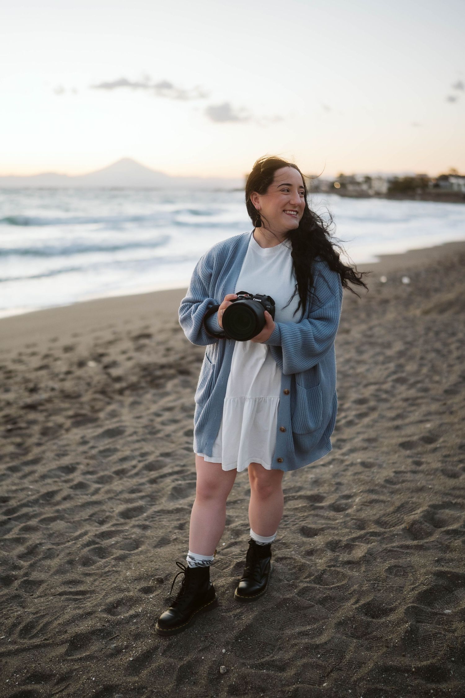 A photographer in a blue cardigan and white dress stands on a beach at sunset holding a camera in coastal setting.