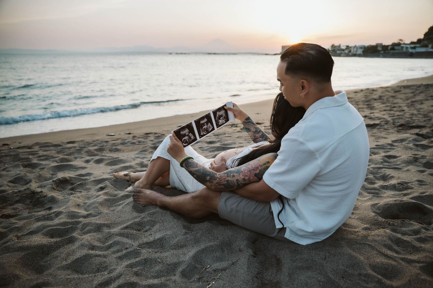 Person in white shirt and gray pants relaxes on sandy beach while reading book at sunset.