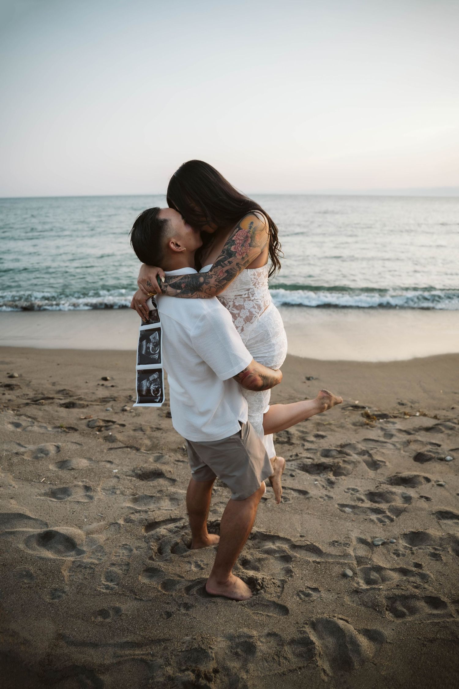 Couple sharing intimate moment on beach while holding ultrasound photo.