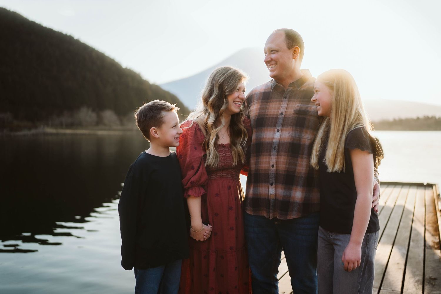 A family stands together on a wooden dock by a serene lake during sunset with mountains in the background.