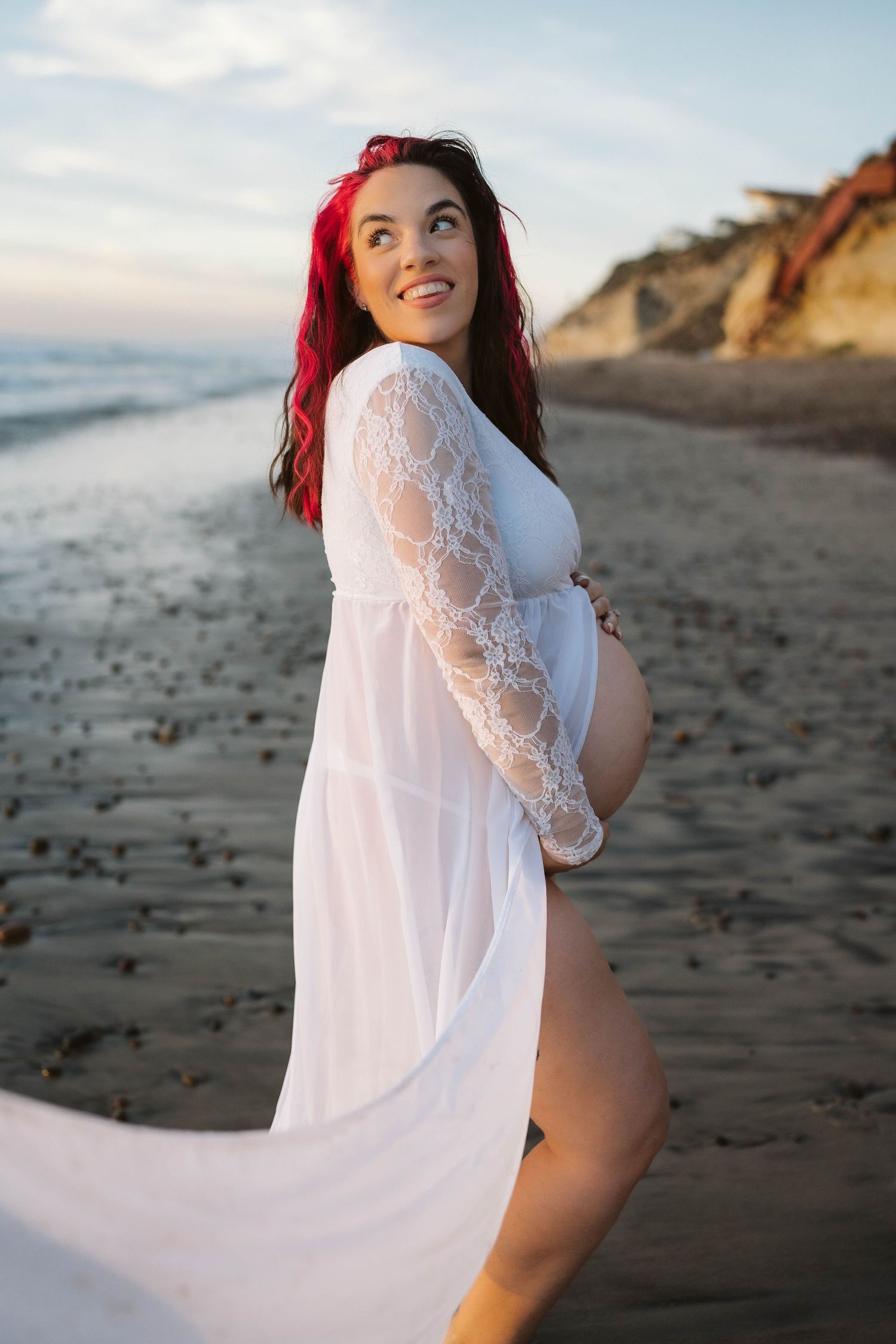A pregnant person in a flowing white lace dress poses on a beach at sunset with cliffs in the background.