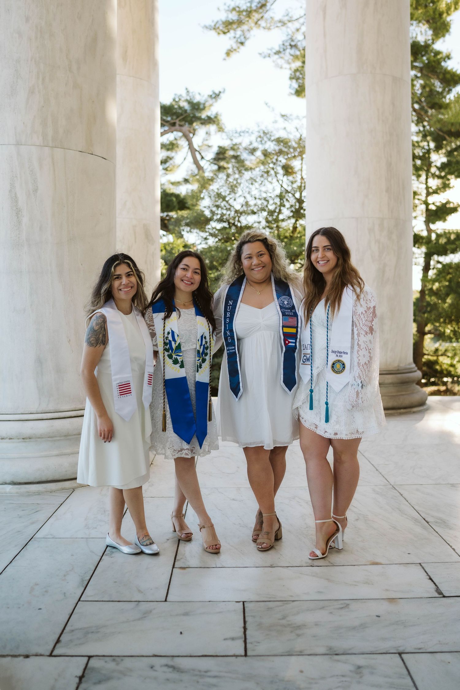 Four graduates wearing graduation sashes and white dresses pose together between classical stone columns on campus.