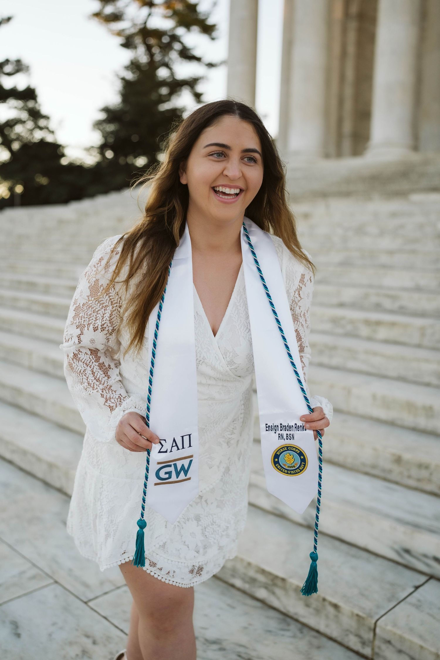 Graduate wearing a white lace dress and graduation stole poses with a big smile on steps of GW University building.
