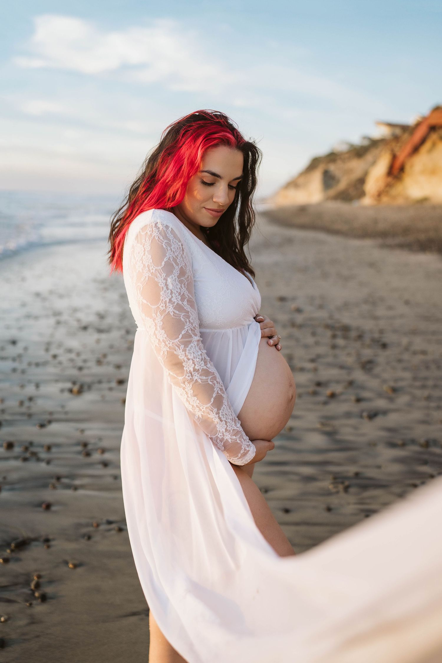 A pregnant woman in a white lace dress stands on a beach at sunset with cliffs in the background.