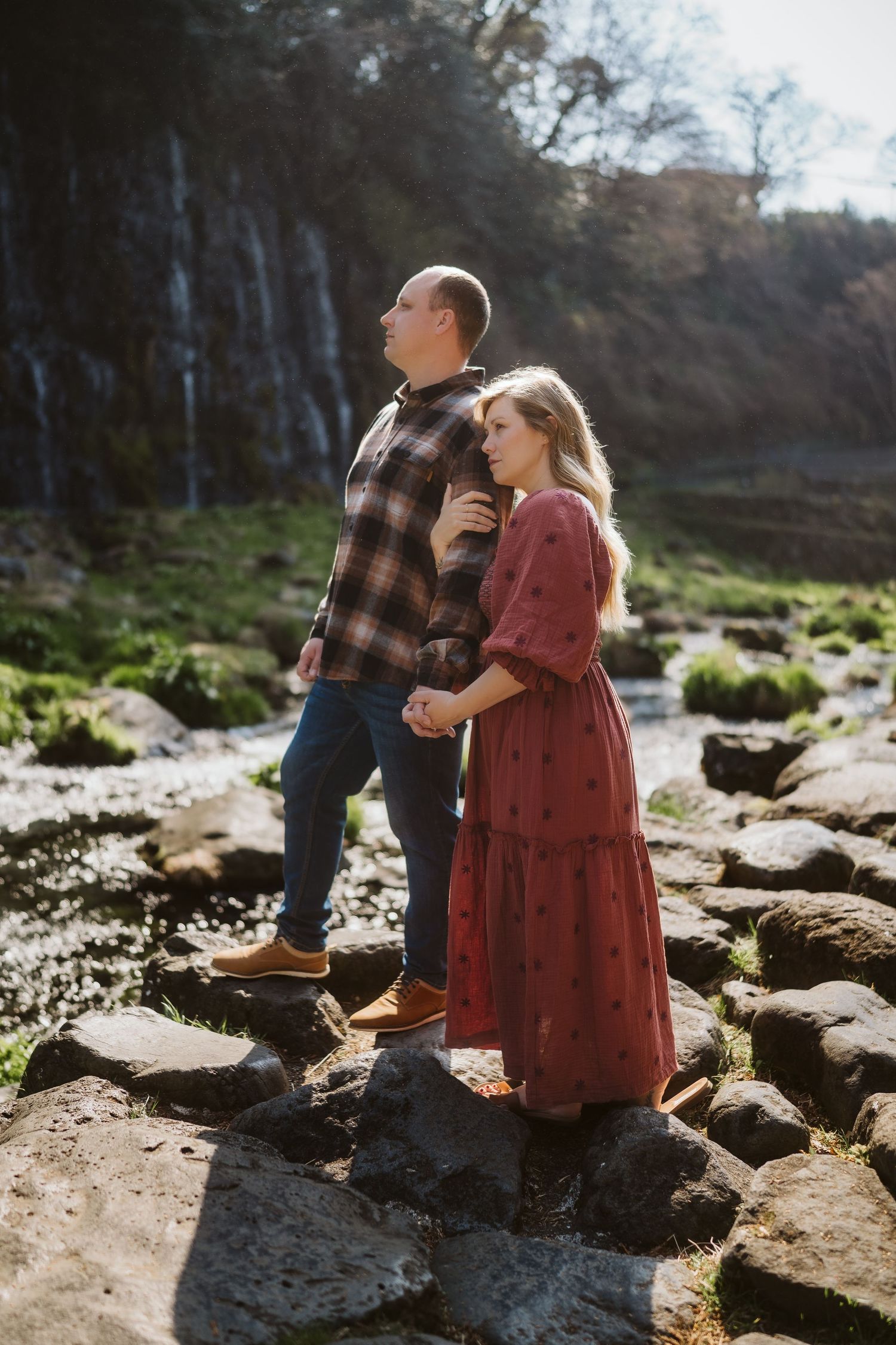 A couple in casual attire stands together on rocky terrain near a stream in soft natural lighting.