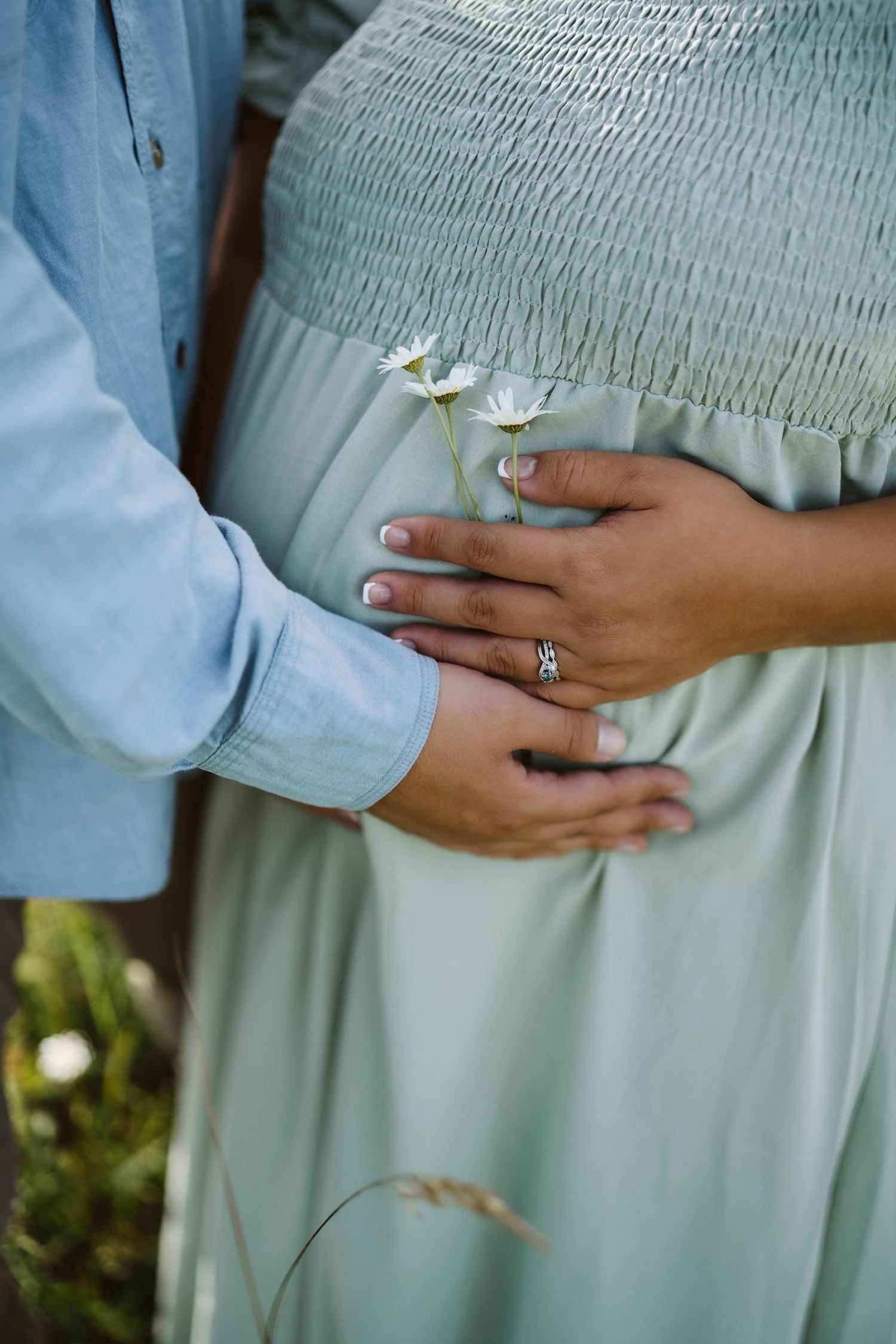 Hands tenderly rest on a sage green smocked maternity dress with a small white floral accent.