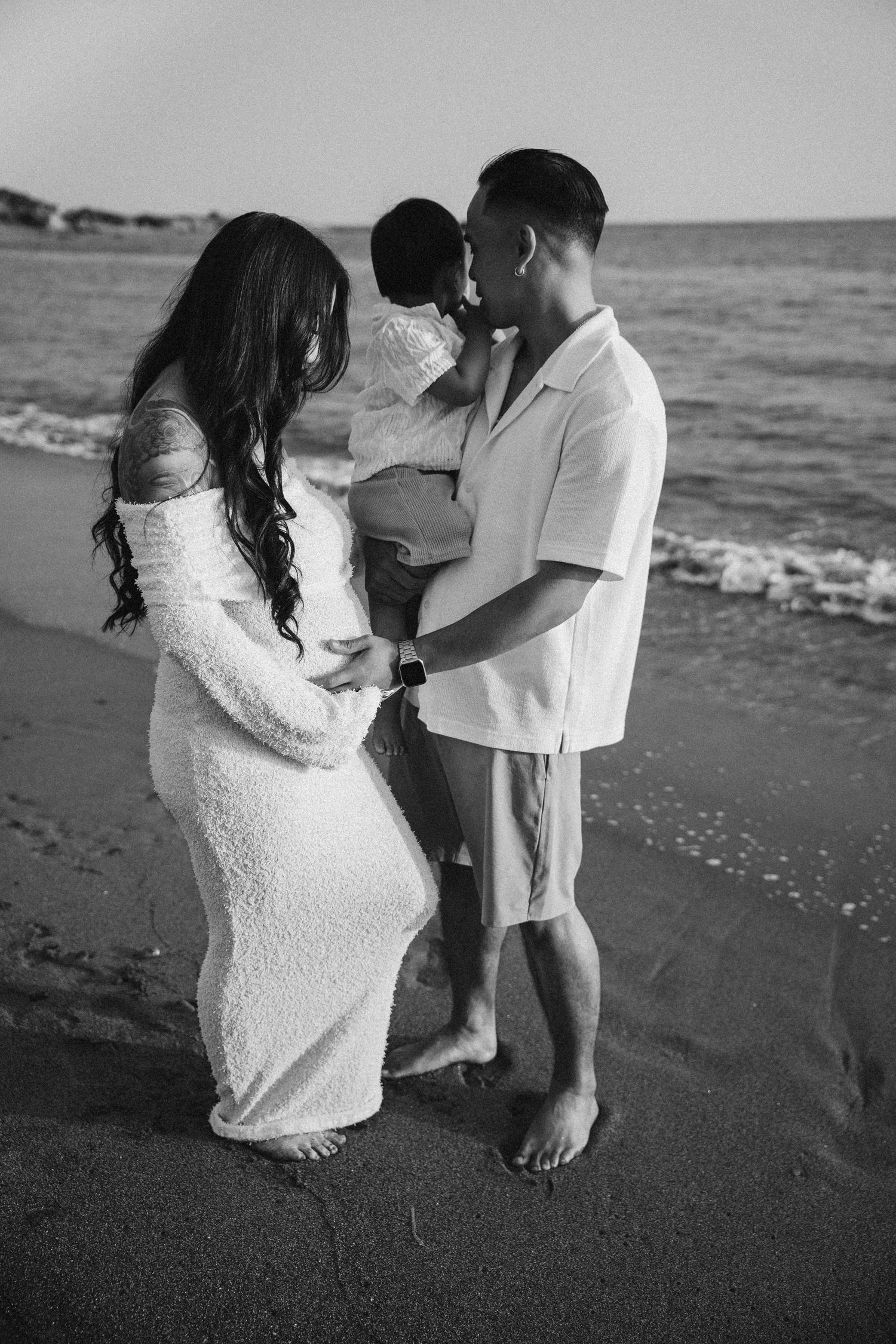 A playful couple in white outfits dances together on a dark sandy beach at sunset.